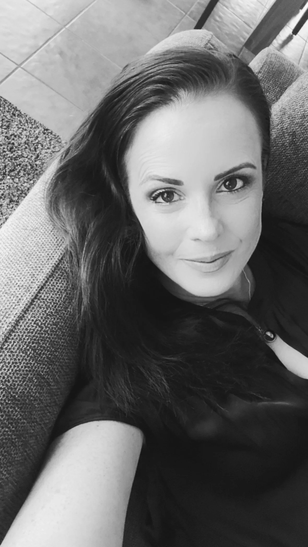 A woman with long dark hair and light skin lying on a couch, looking up at the camera, in grayscale.