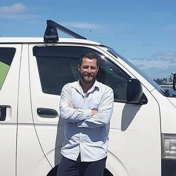 Tom the owner- a friendly man with beard in white shirt standing with arms crossed in front of a white van on a sunny day.