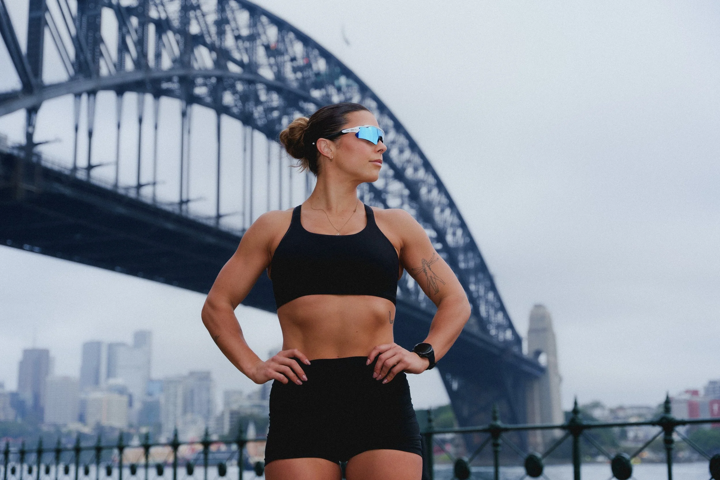 A fit woman with a tattoo on her left arm posing outdoors near a river with a large bridge in the background. She is wearing sunglasses, a black sports bra, black shorts, and a fitness tracker on her left wrist.