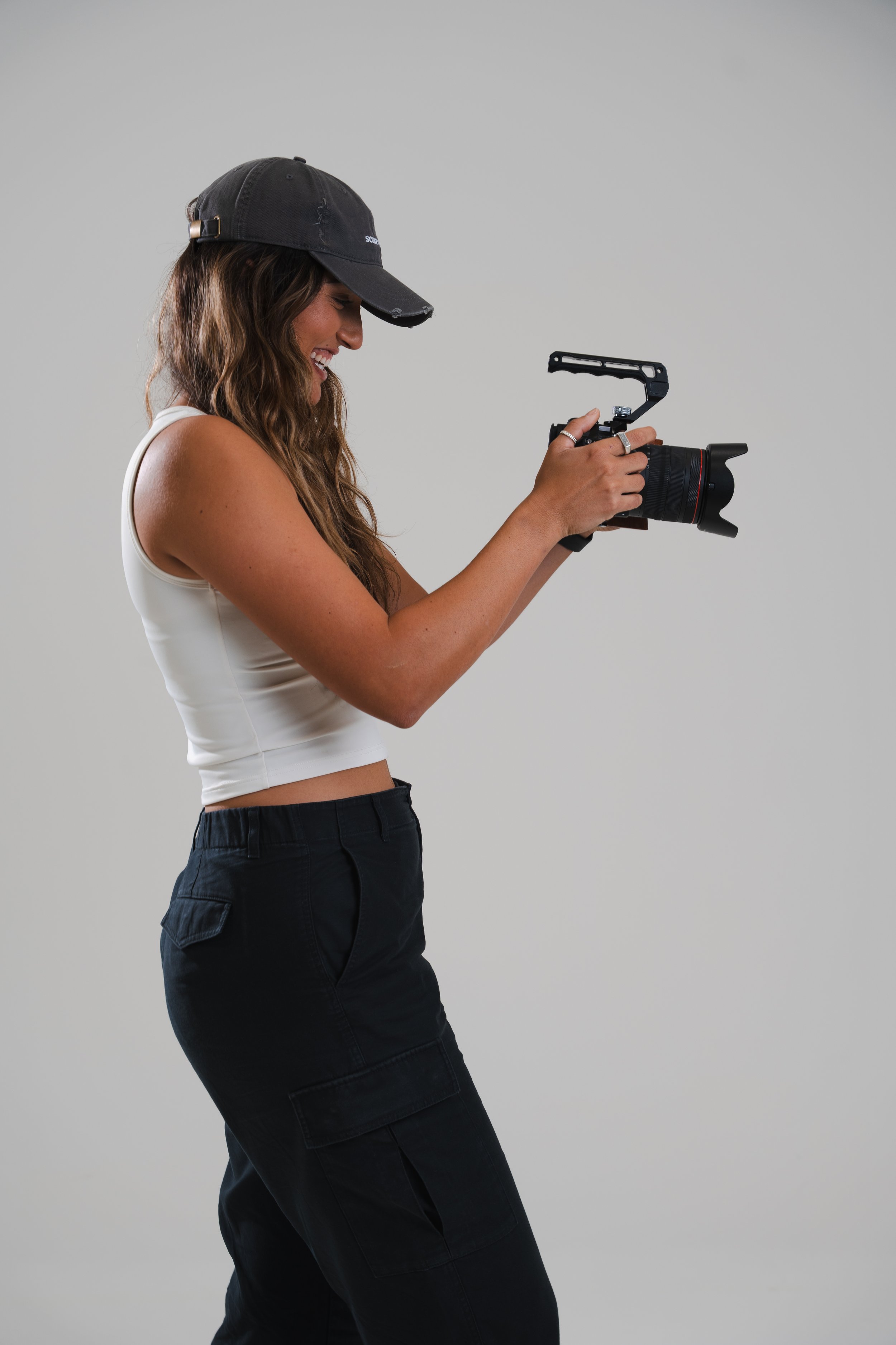 Woman with long brown hair wearing a black cap, white tank top, and black cargo pants, holding a camera with a stabilizer rig, smiling while looking at the camera.