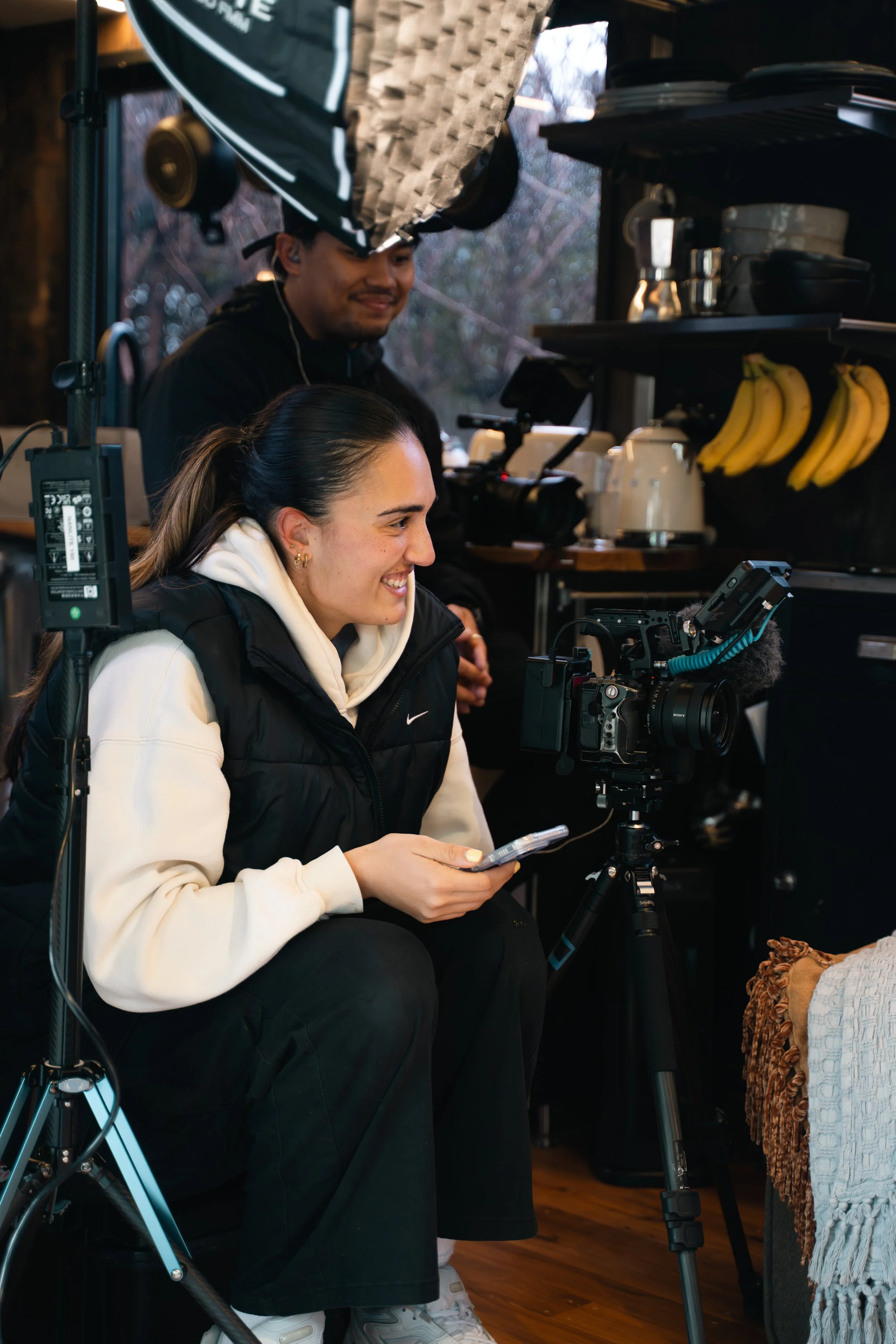 Two people working with video equipment inside a cozy room with a window, bananas hanging on a shelf, and various kitchen items.