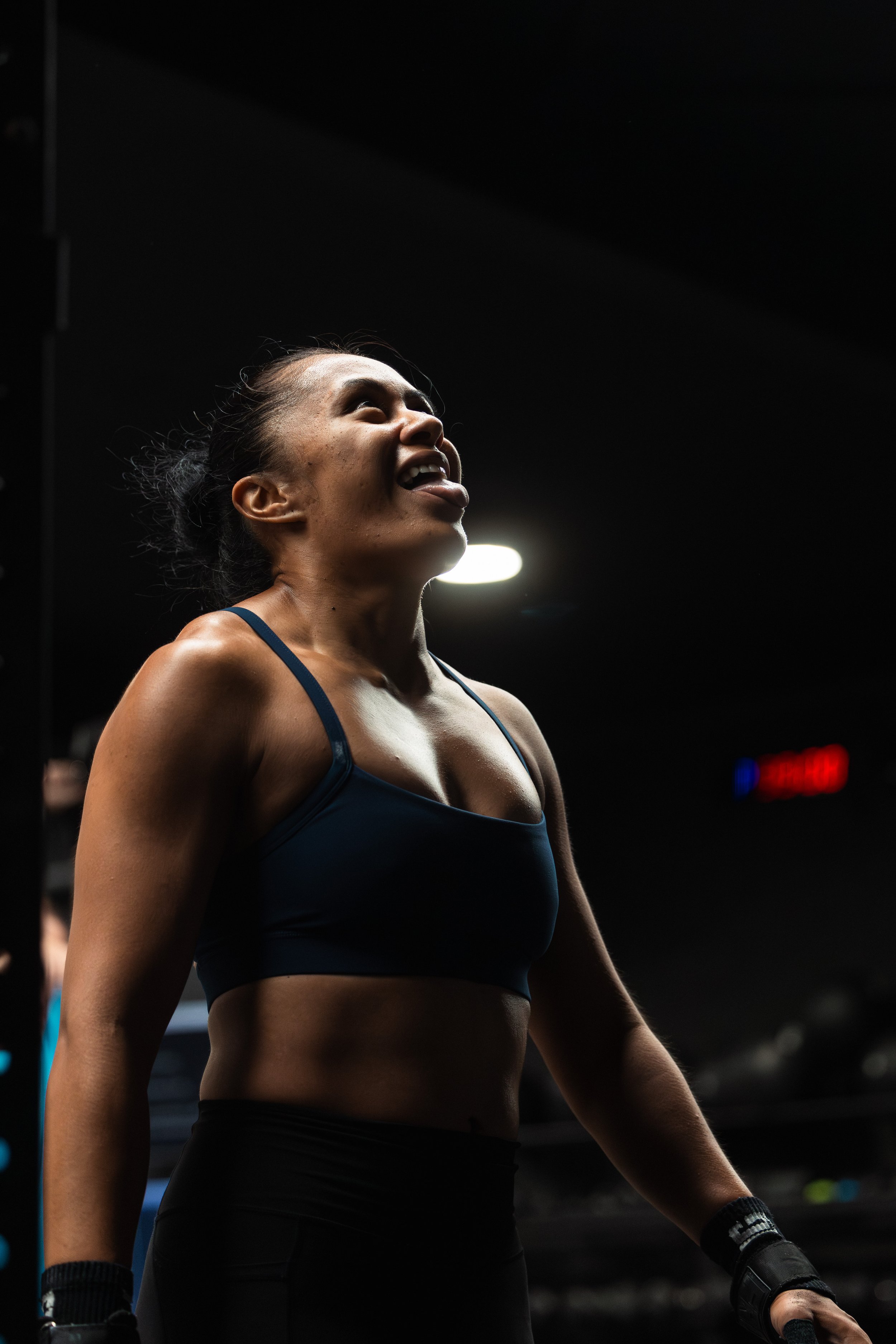 A female athlete with sweat on her face, wearing a dark blue sports bra and black workout gloves, standing in a gym with a dark background and bright lights.
