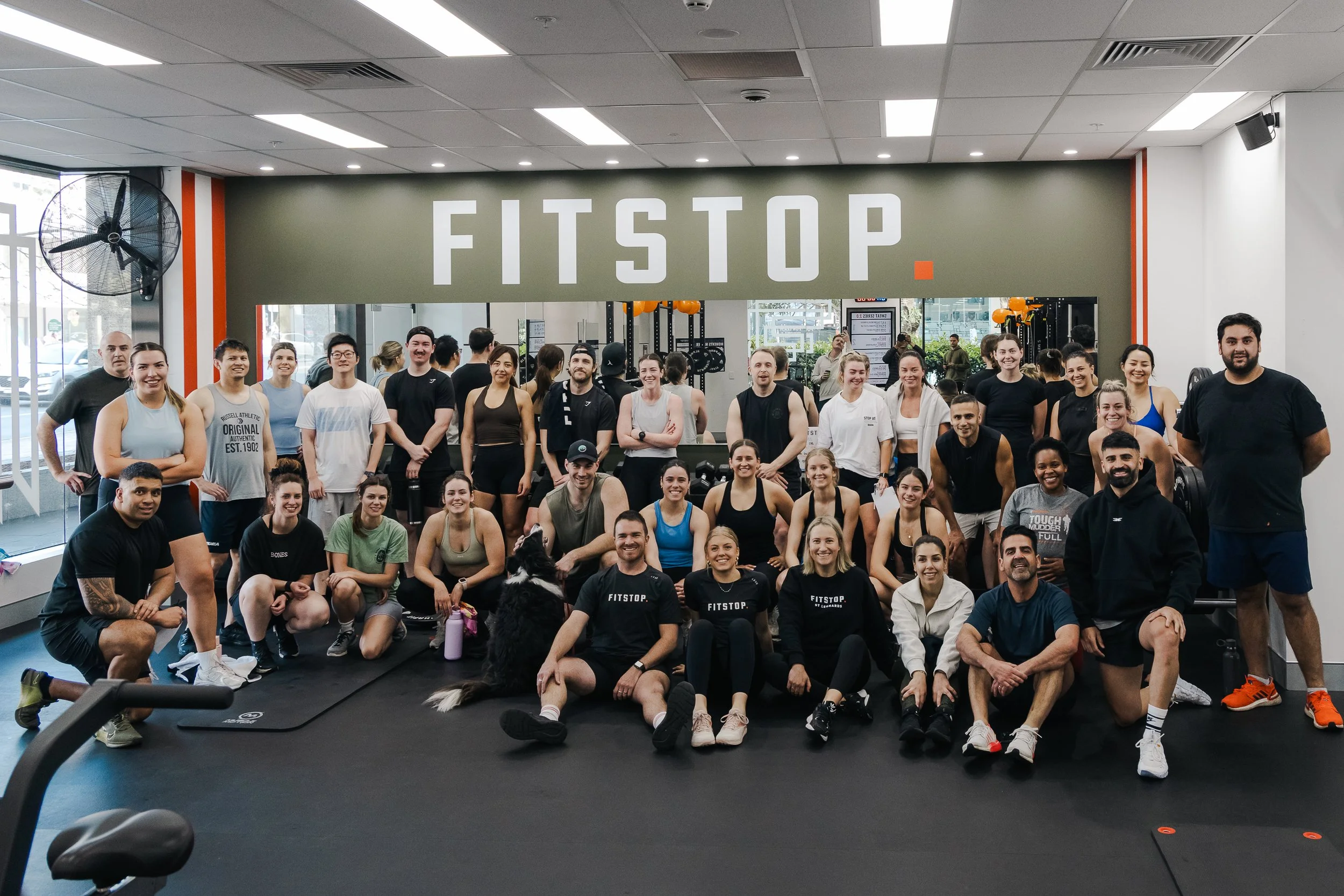 Group photo of a large diverse group of people in a fitness gym, smiling and posing together, with a gray wall behind them displaying the word 'FISTOP' in large white letters.