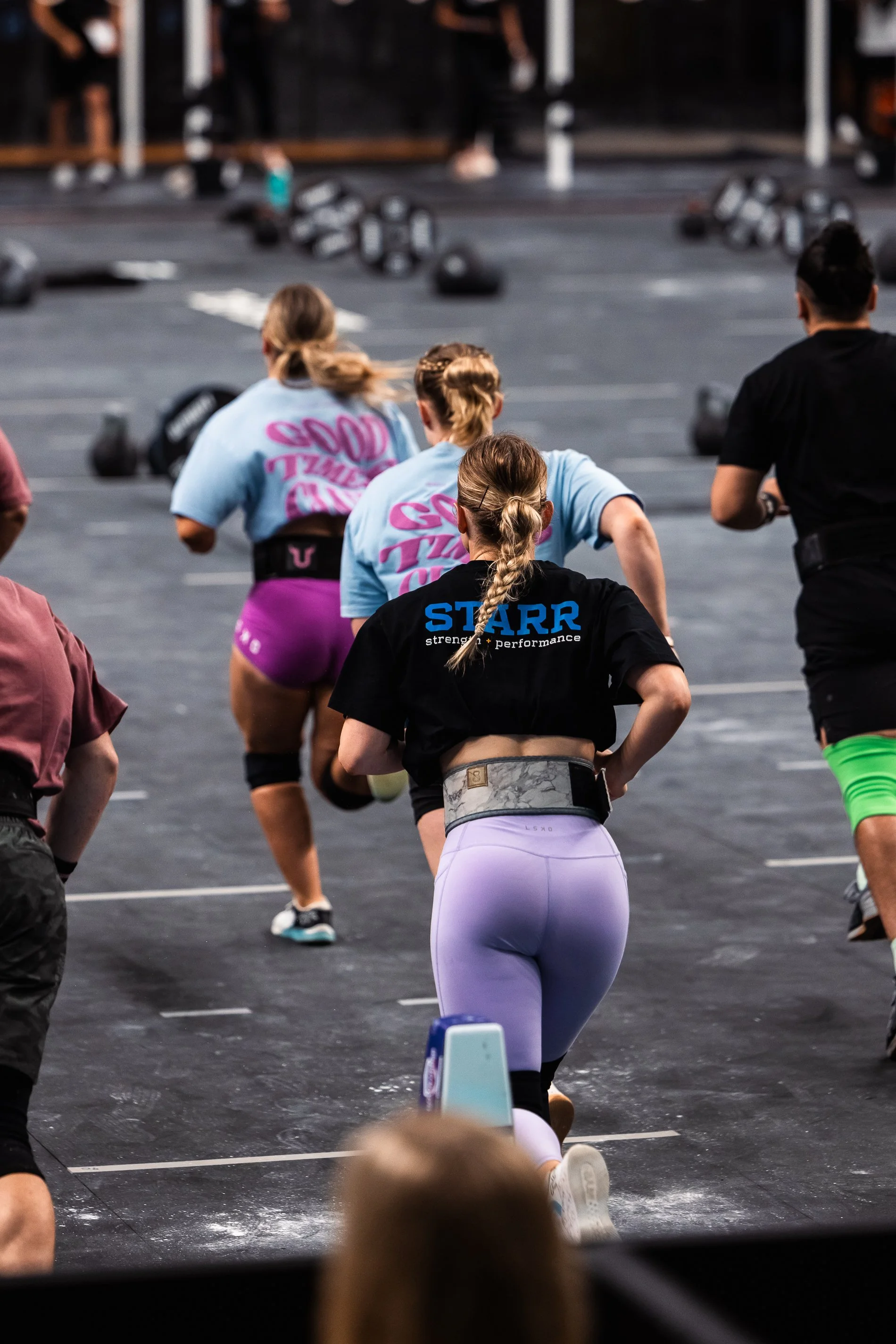 Group of people participating in a fitness class, with kettlebells on the floor and a woman kneeling in the foreground, wearing athletic wear and a weightlifting belt.