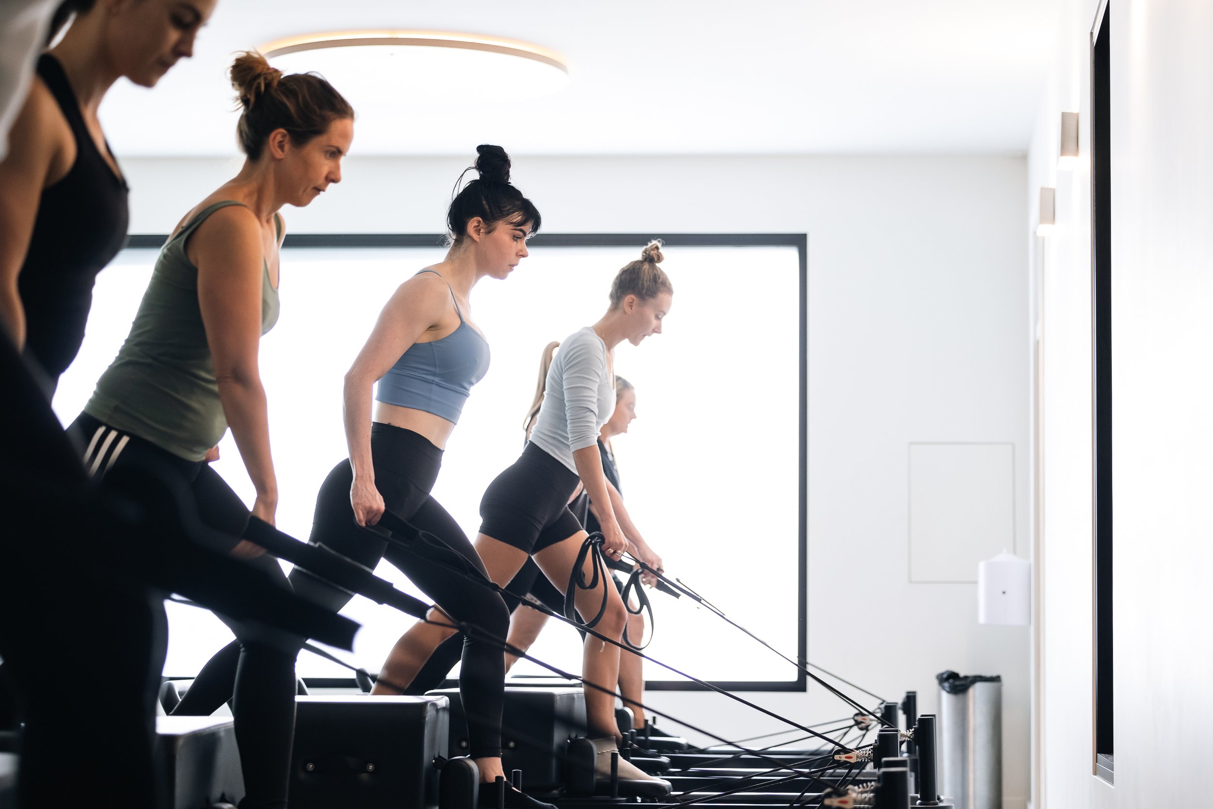 Five women using Pilates reformer machines in a bright fitness studio.