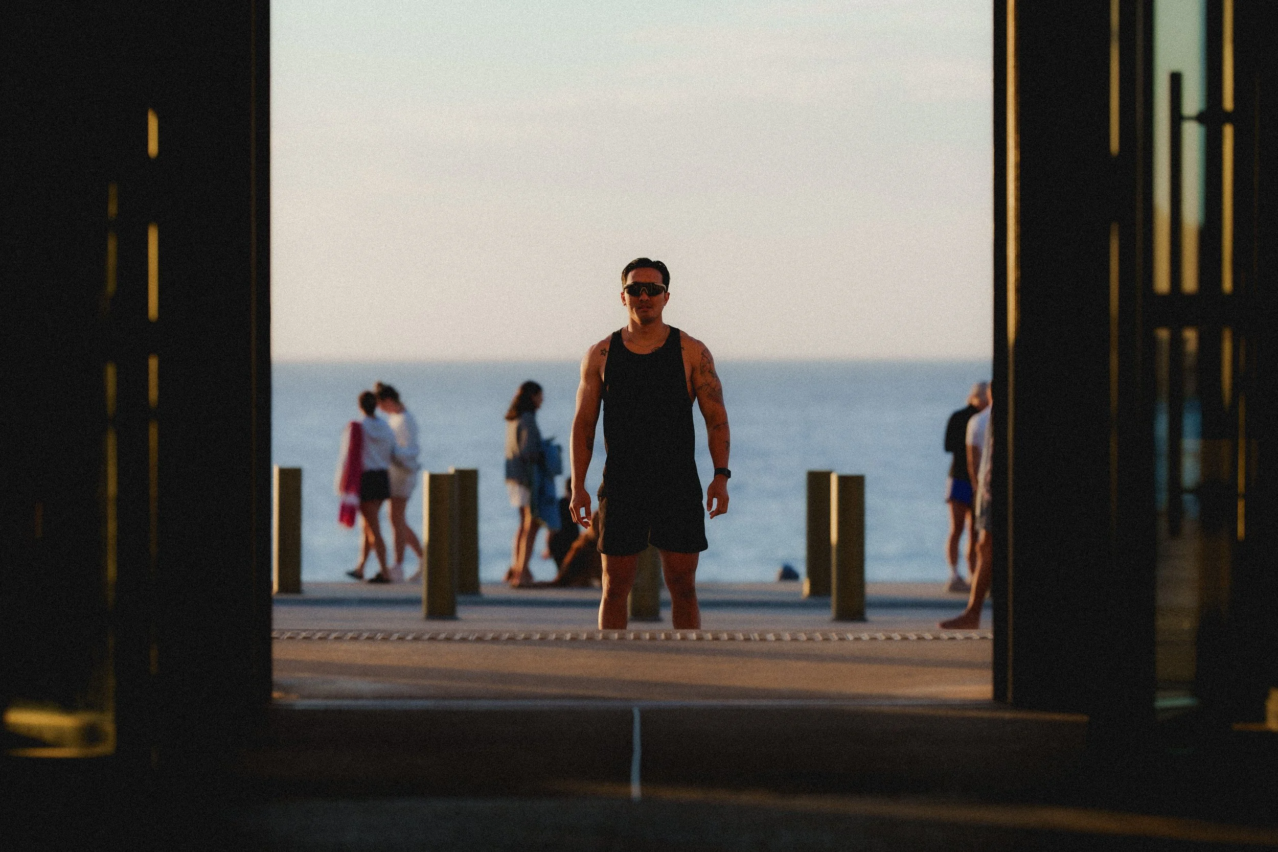 A man in black tank top and shorts standing on a pier with ocean in background, viewed from inside a dark doorway, with people walking along the pier.