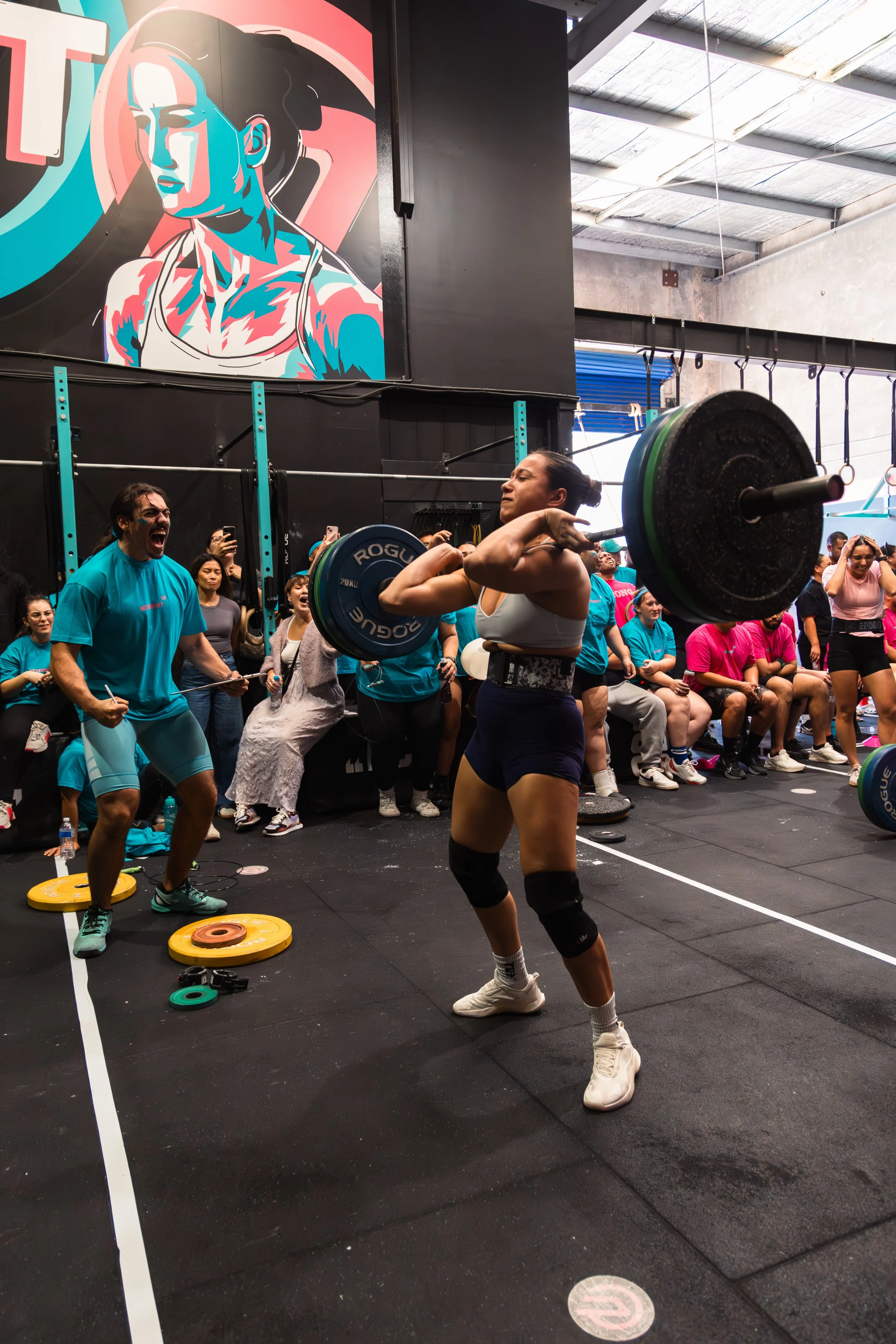 A woman performs a clean and jerk weightlifting move in a gym with an audience watching. She wears a white sports bra, black shorts, knee wraps, and white athletic shoes. The gym has a black floor, with a large colorful mural of a woman lifting weights on the wall, and spectators seated along the side.