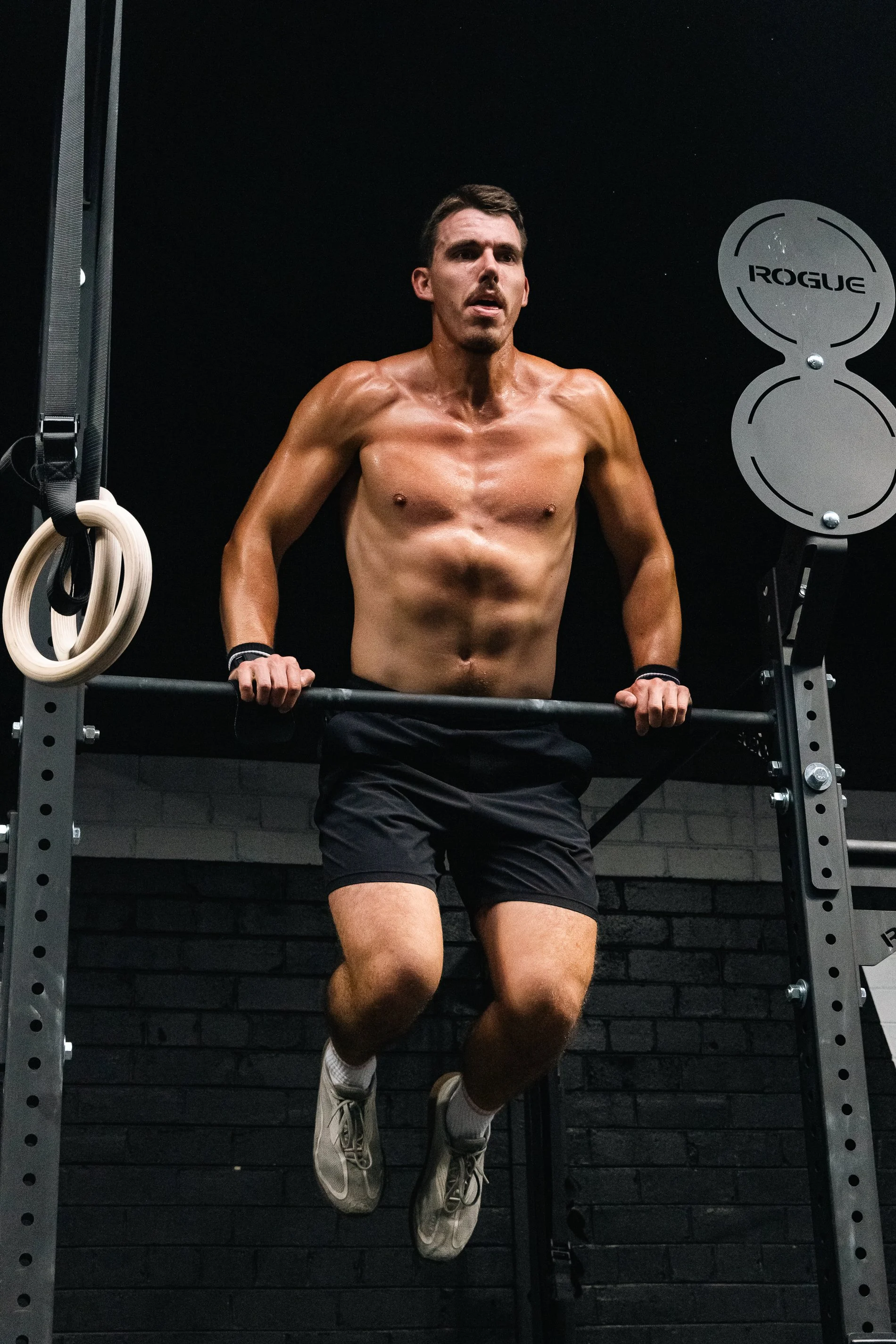 A shirtless man with dark shorts and sneakers performing a pull-up in a gym.