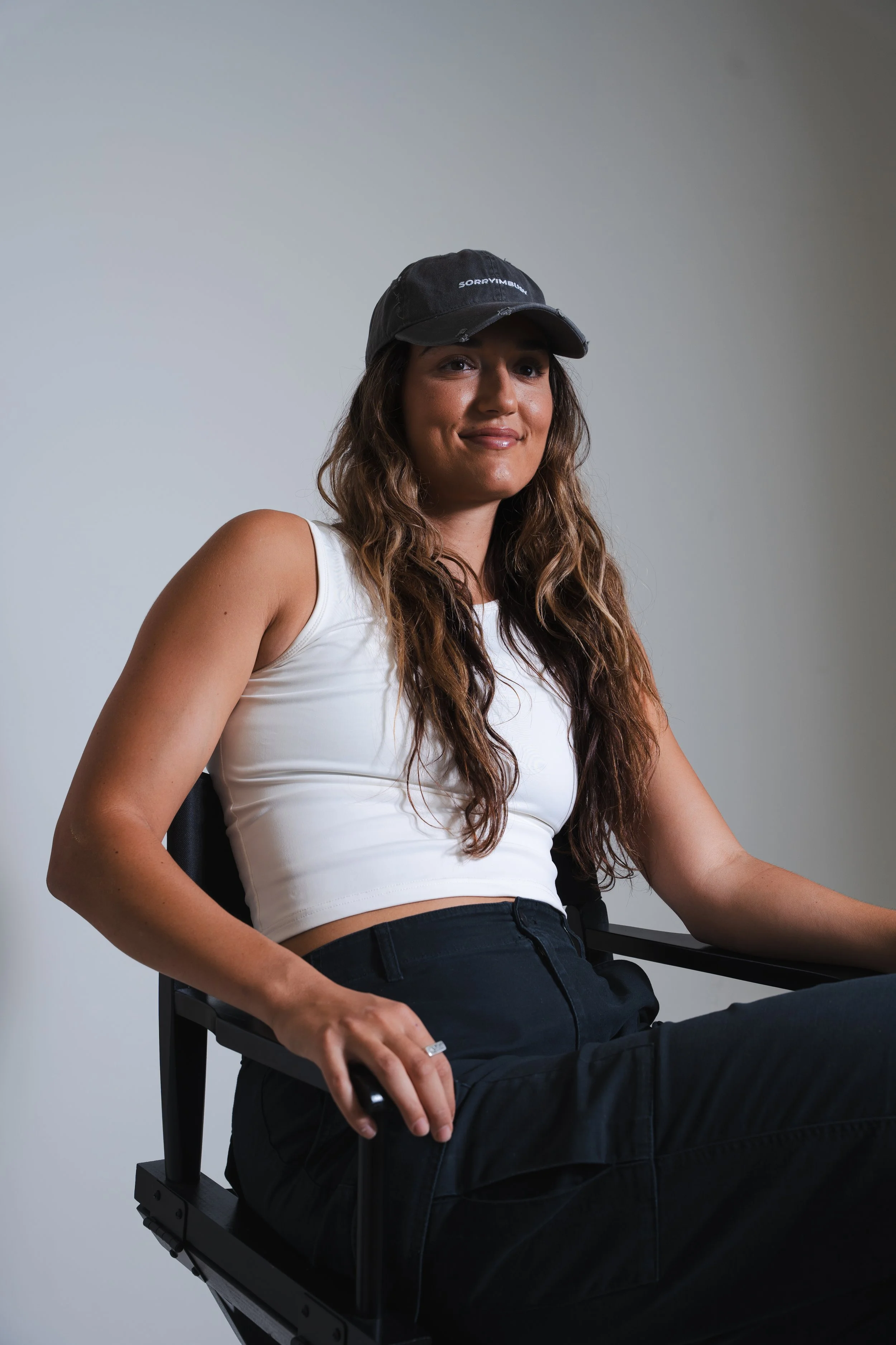 A woman with long wavy brown hair, wearing a black cap and white tank top, sitting on a black chair against a plain light gray background.
