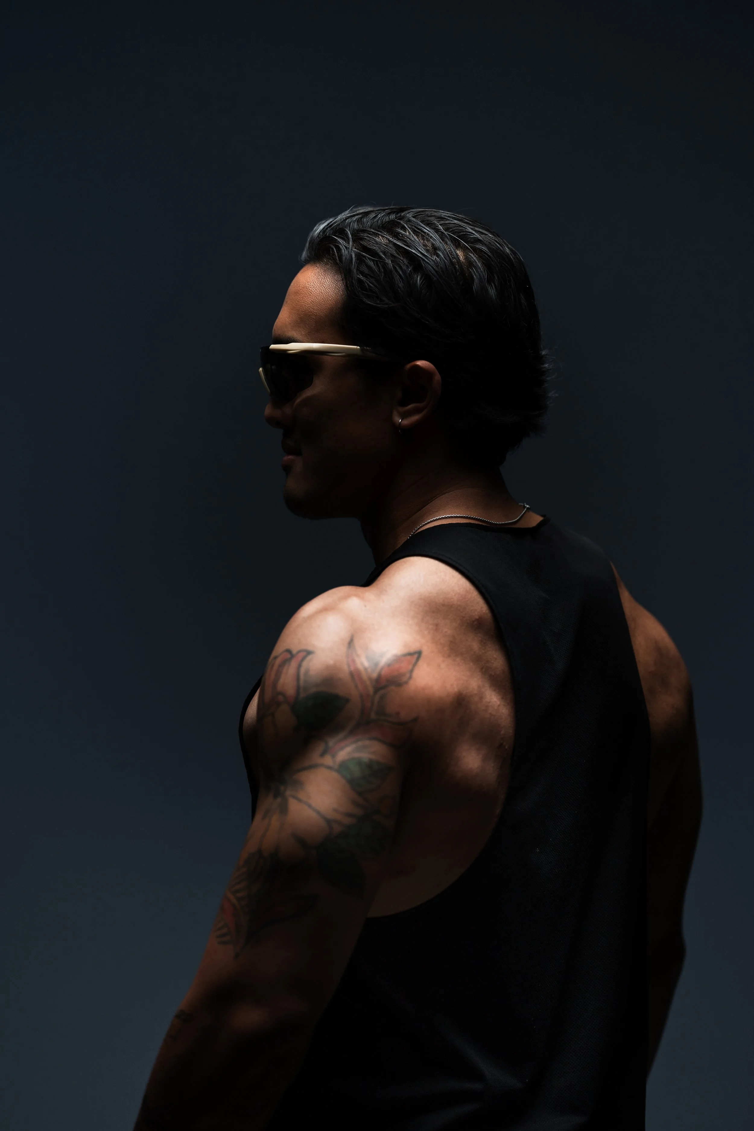 Side profile of a dark-haired man wearing sunglasses and a black sleeveless shirt, showing a tattooed arm, against a dark background.