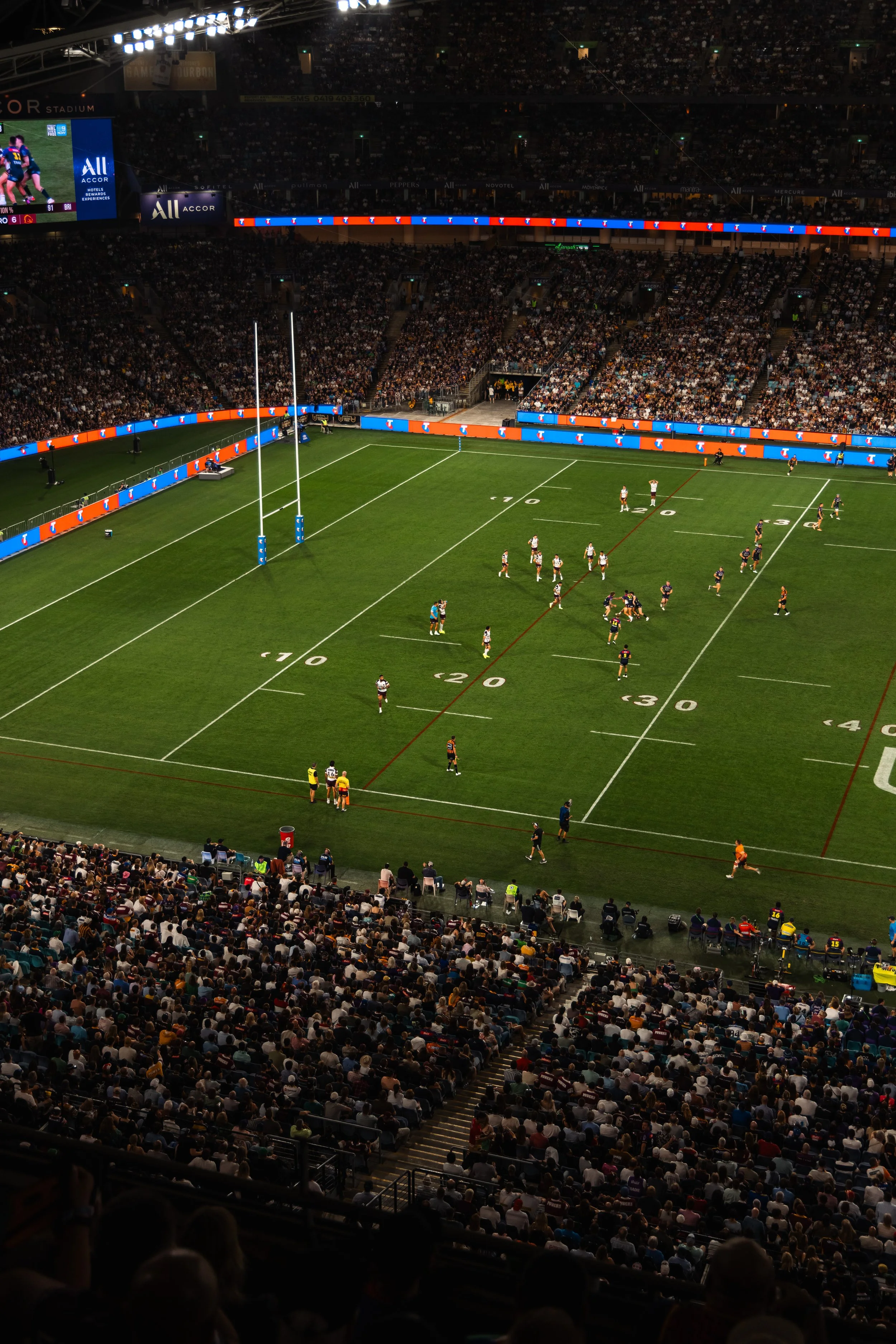 Indoor rugby stadium filled with spectators, with players on the field preparing for a game, field markings visible, large screens showing game action, and bright stadium lighting.