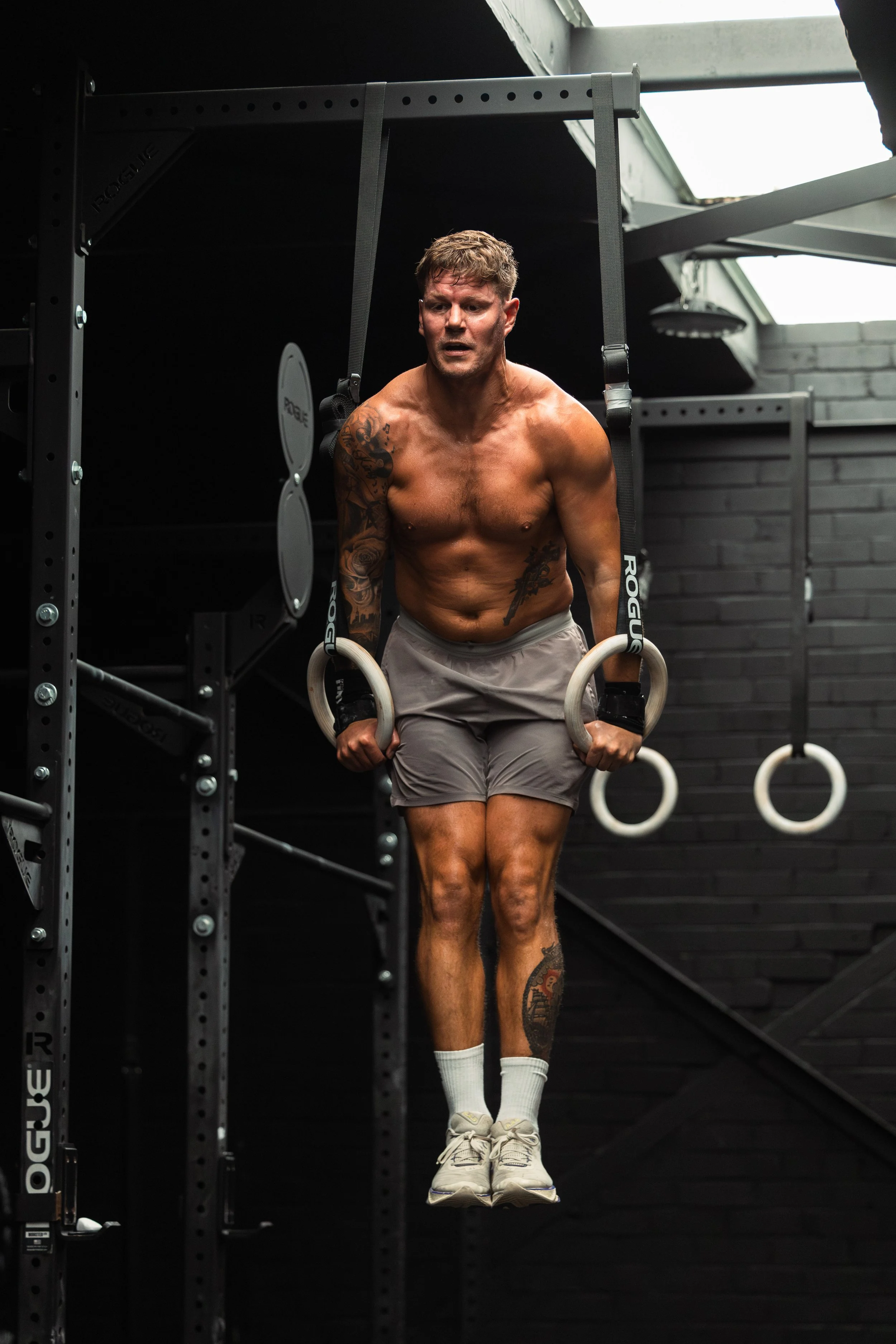 A muscular, shirtless man with tattoos on his arms and leg, wearing gray shorts, white socks, and sneakers, is performing a gymnastics exercise on gymnastic rings in a black indoor gym.