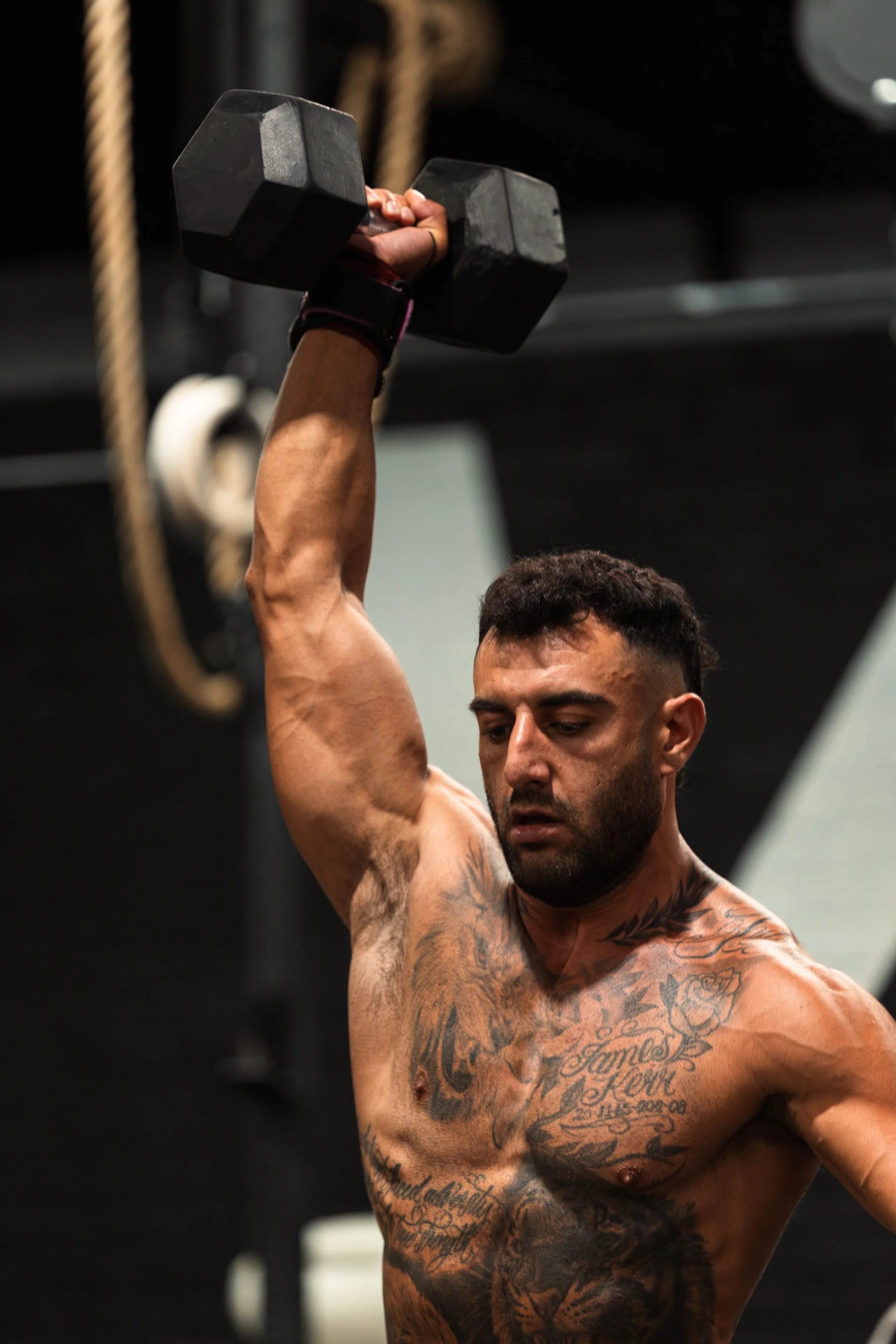 A shirtless man with tattoos lifting a black dumbbell overhead during a workout in a gym.