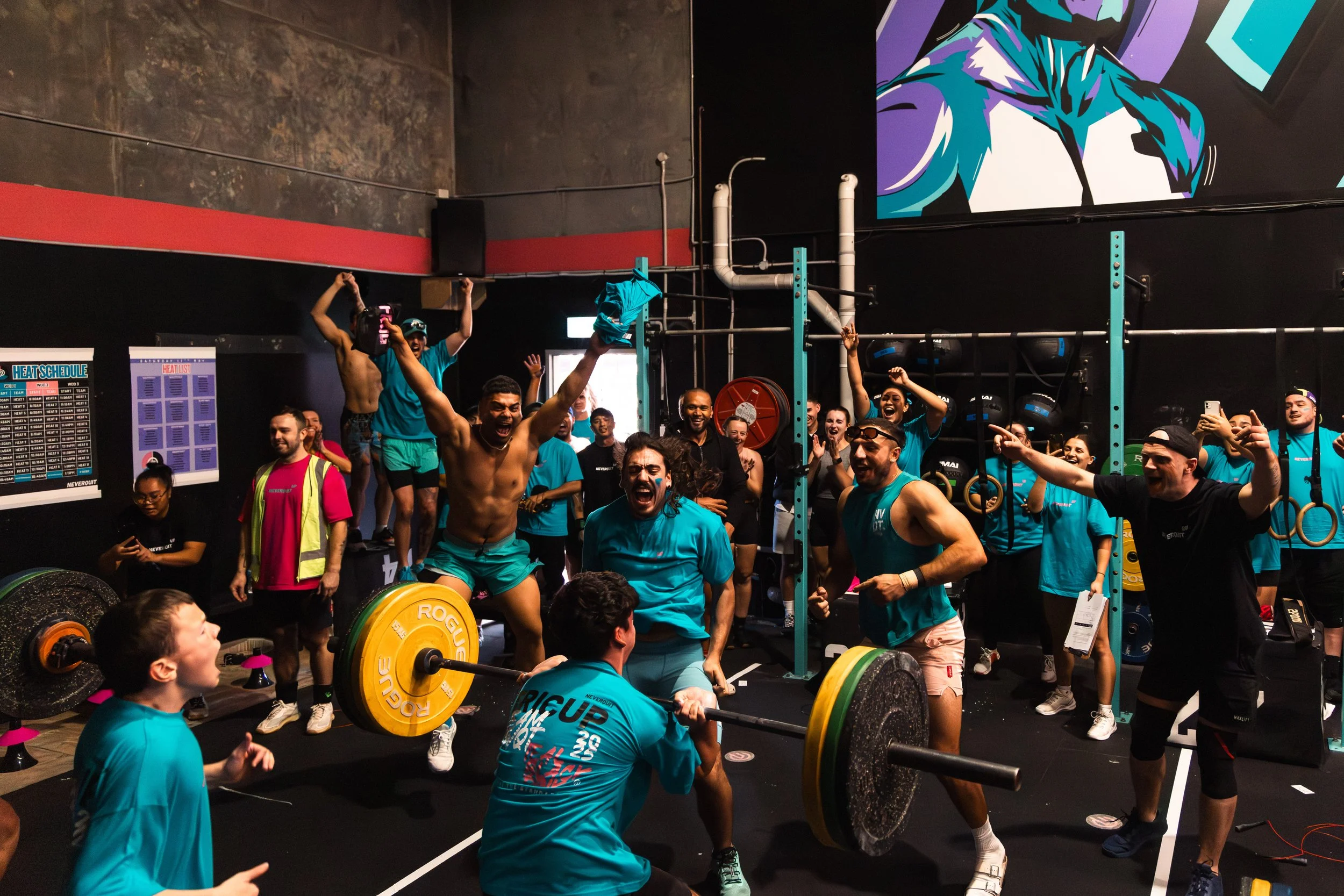Group of people celebrating after a weightlifting competition, with one person lifting a barbell overhead, in a gym with black walls, colorful posters, and gym equipment.
