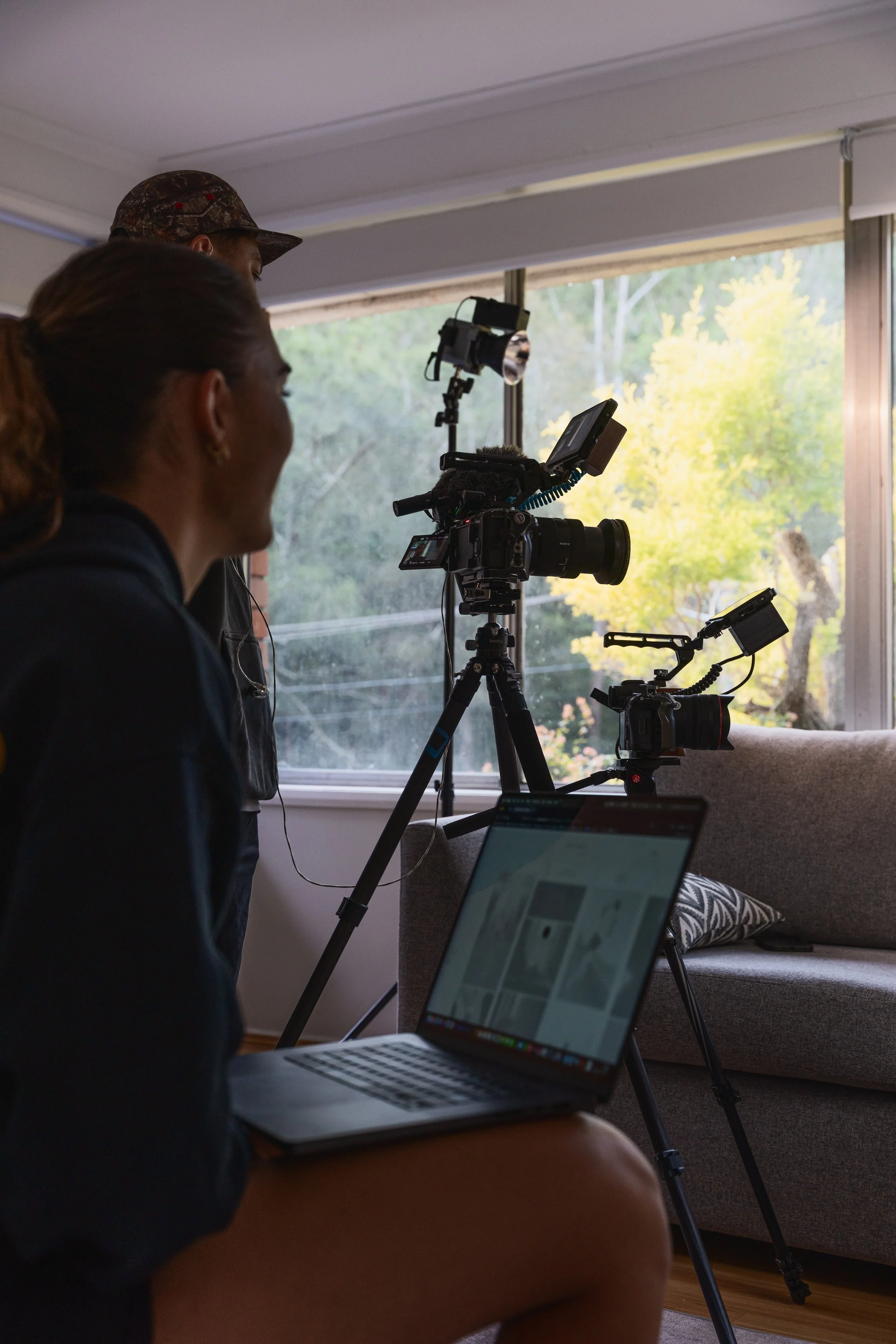 Two people working with cameras set up on tripods inside a room with large windows showing trees outside.