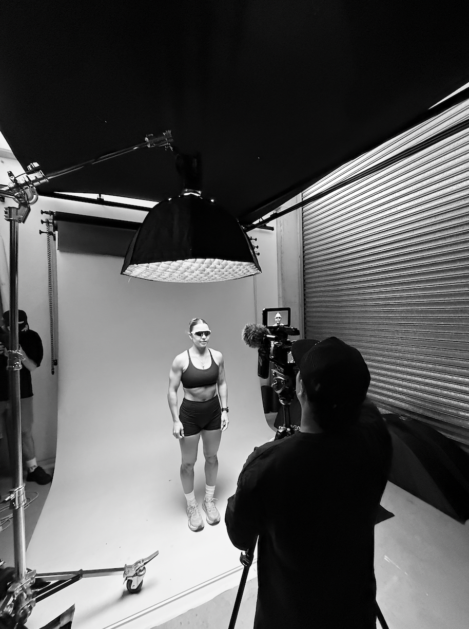 A woman in athletic wear and sunglasses standing in front of a camera in a photo studio with a plain backdrop.