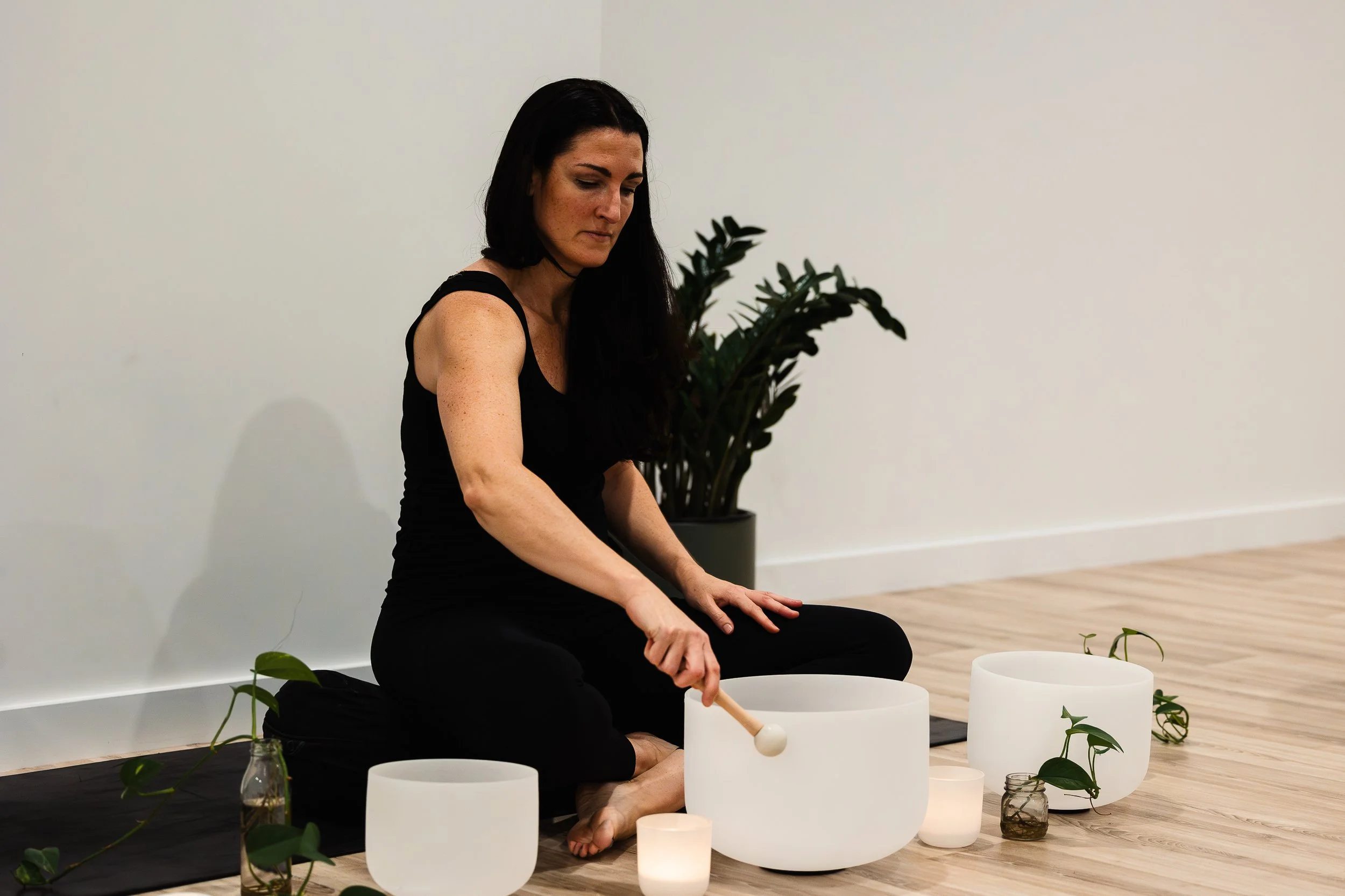 Woman sitting cross-legged on the floor playing crystal singing bowls, surrounded by candles and plants, in a minimalist room with a wooden floor and white walls.