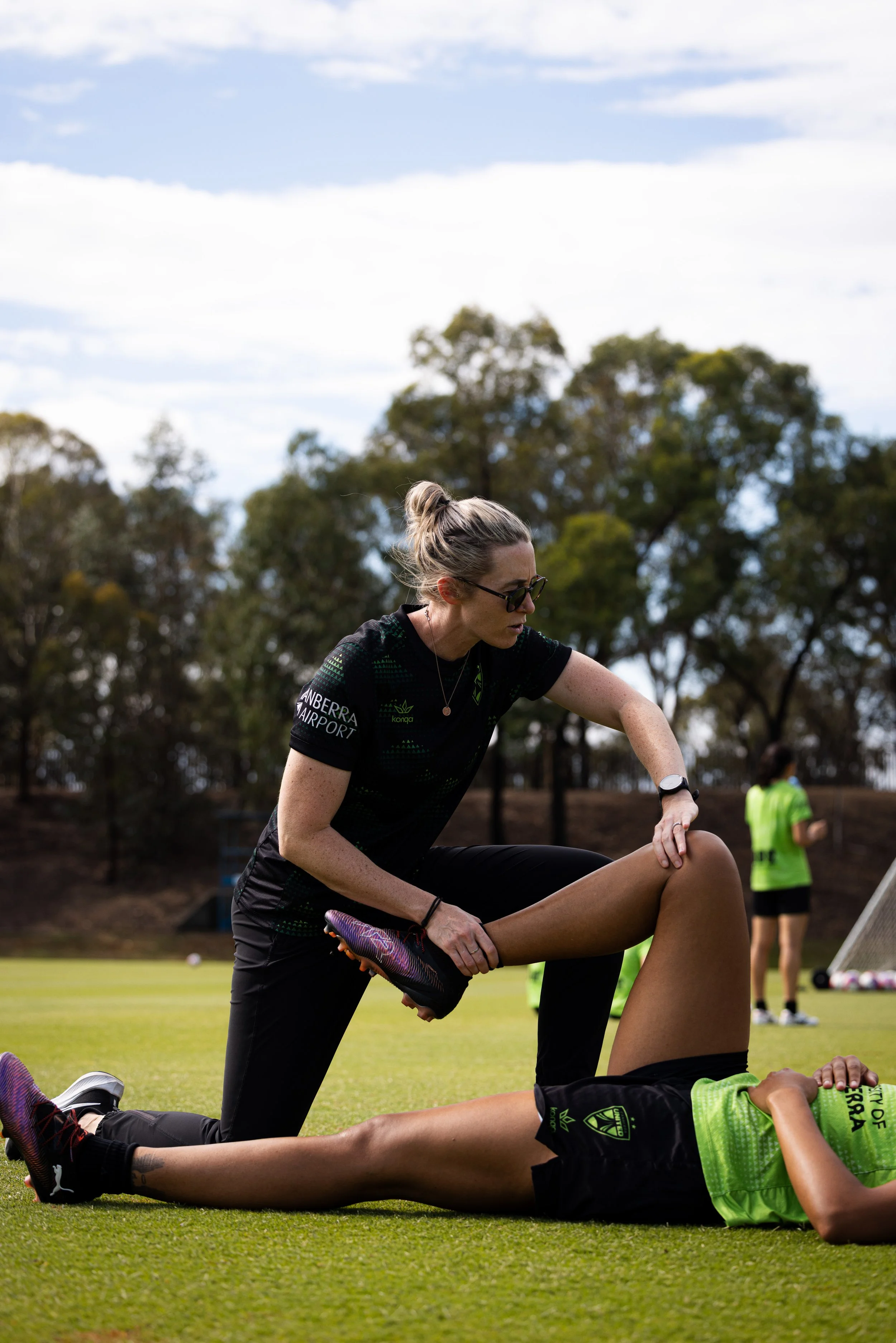 A woman in black sportswear stretching a soccer player's leg on a green field during practice, with trees and another person in a yellow bib standing in the background.