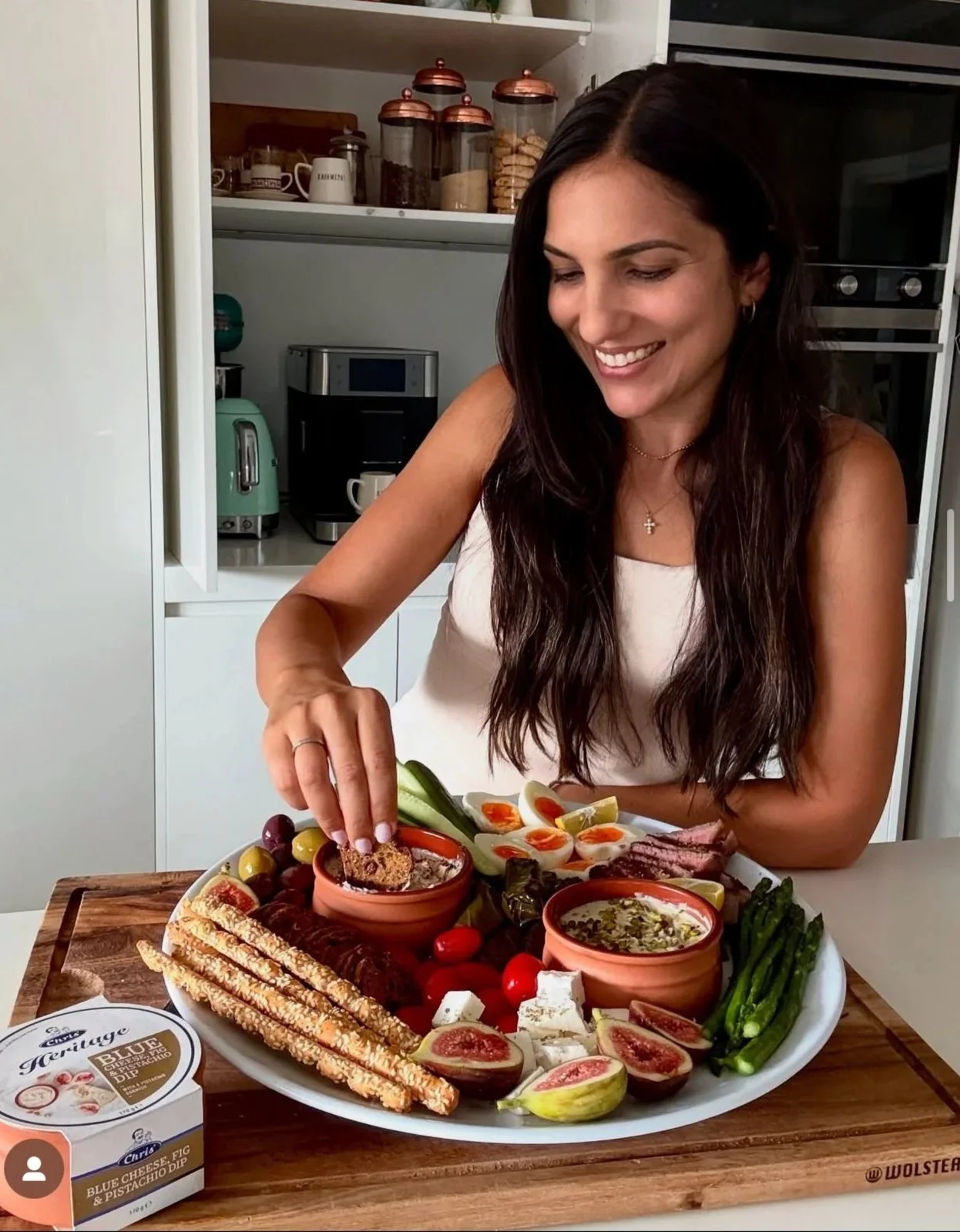 A woman smiling and preparing a charcuterie board with various cheeses, figs, breadsticks, vegetables, and dips in a kitchen.