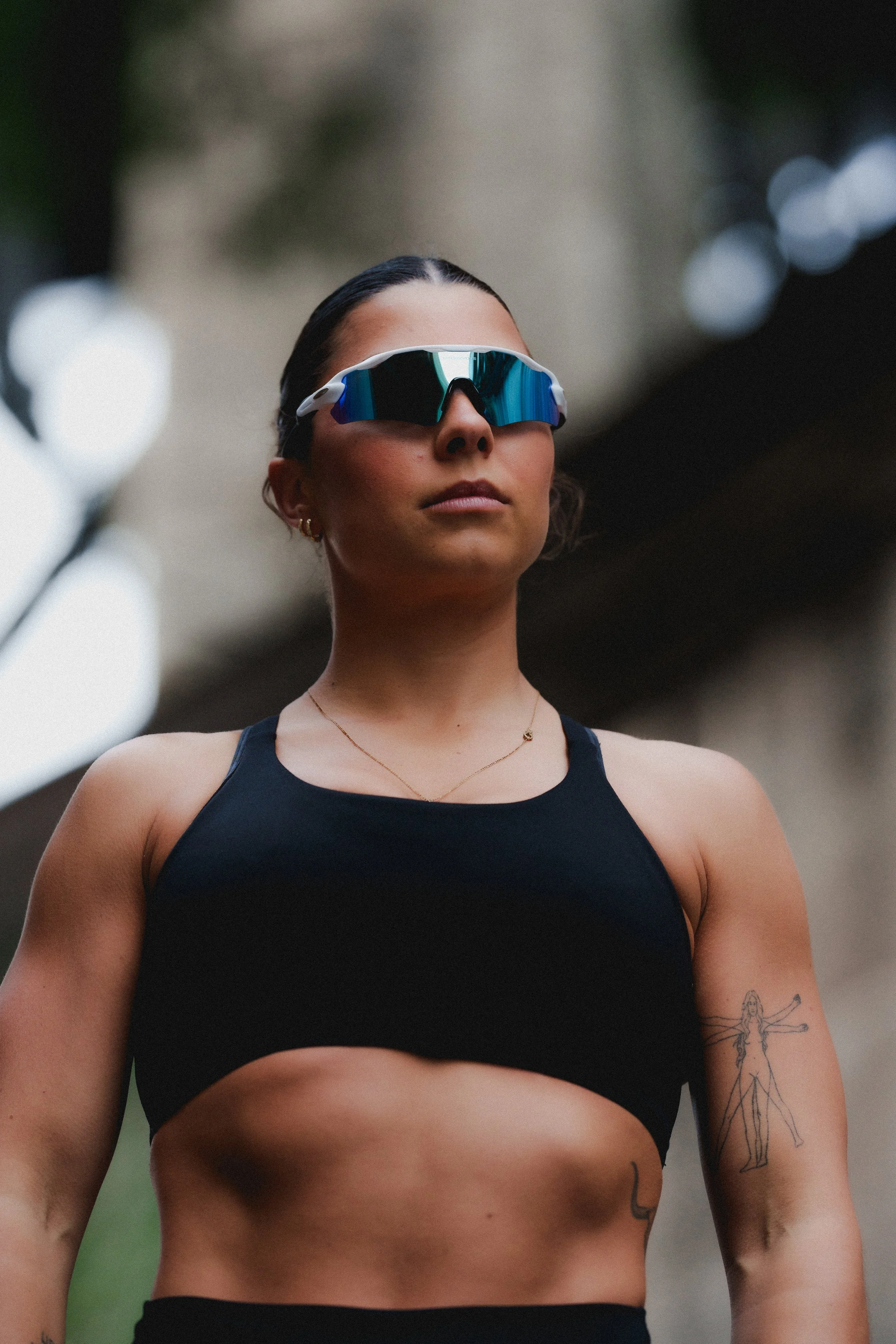 A woman wearing a black sports bra, sunglasses, and a necklace, standing outdoors with blurry trees in the background.