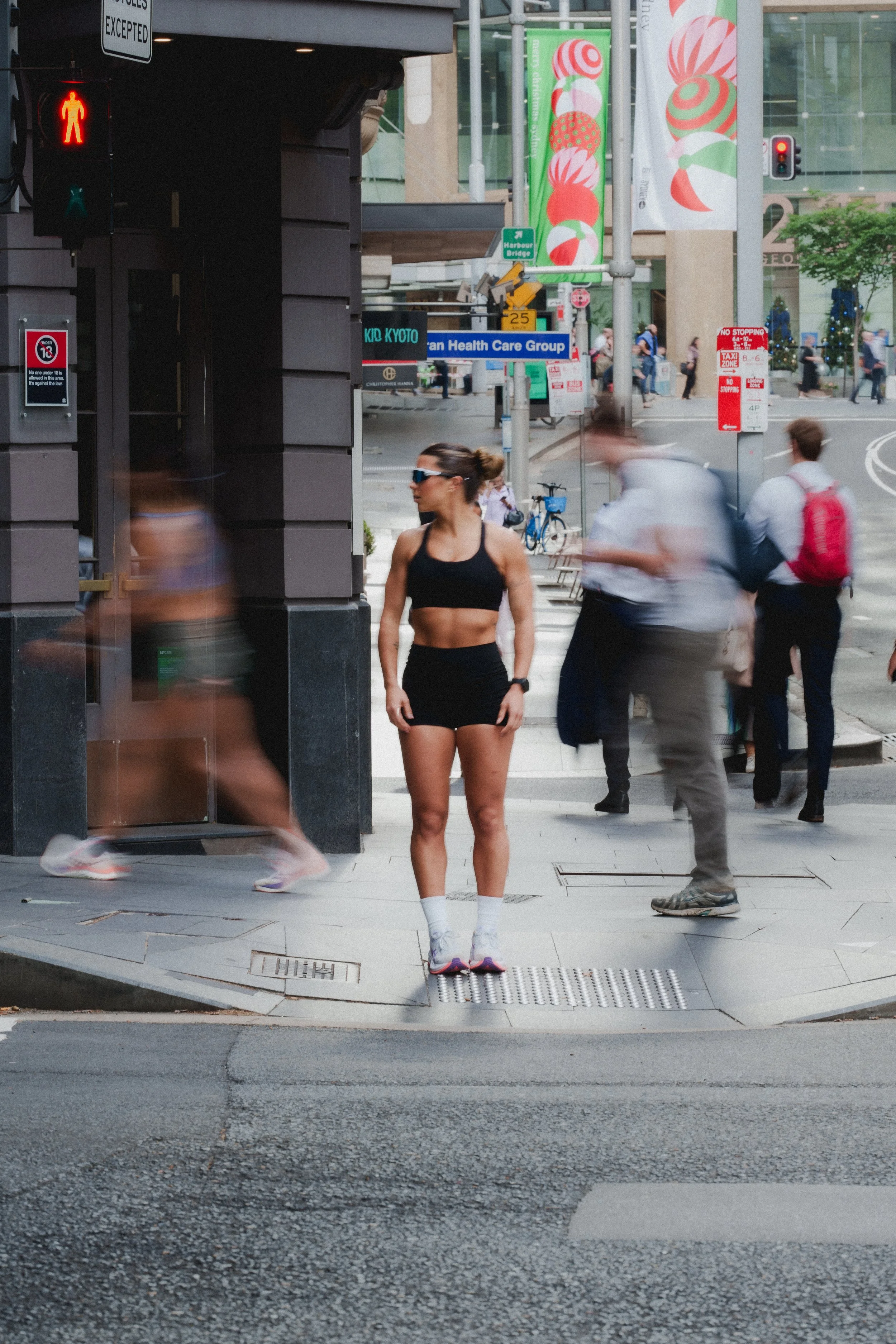 A woman in athletic attire standing at a busy city street corner while other pedestrians walk by in motion blur.