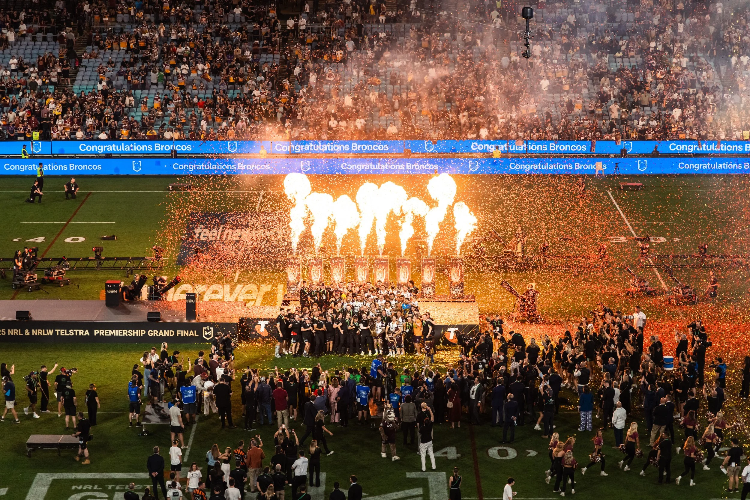 Celebration on a football field with fireworks, confetti, and a large crowd. A team of players and staff stand together while fans gather around. A banner reads 'NRL & NRLW Telstra Premiership Grand Final.'