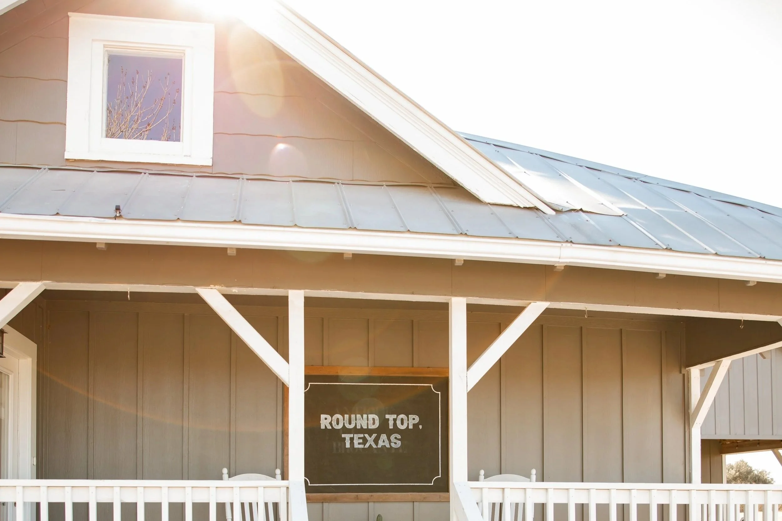 The front of a house with a grey metal roof, a small window at the top, and a porch with a blackboard sign that reads "Round Top, Texas."
