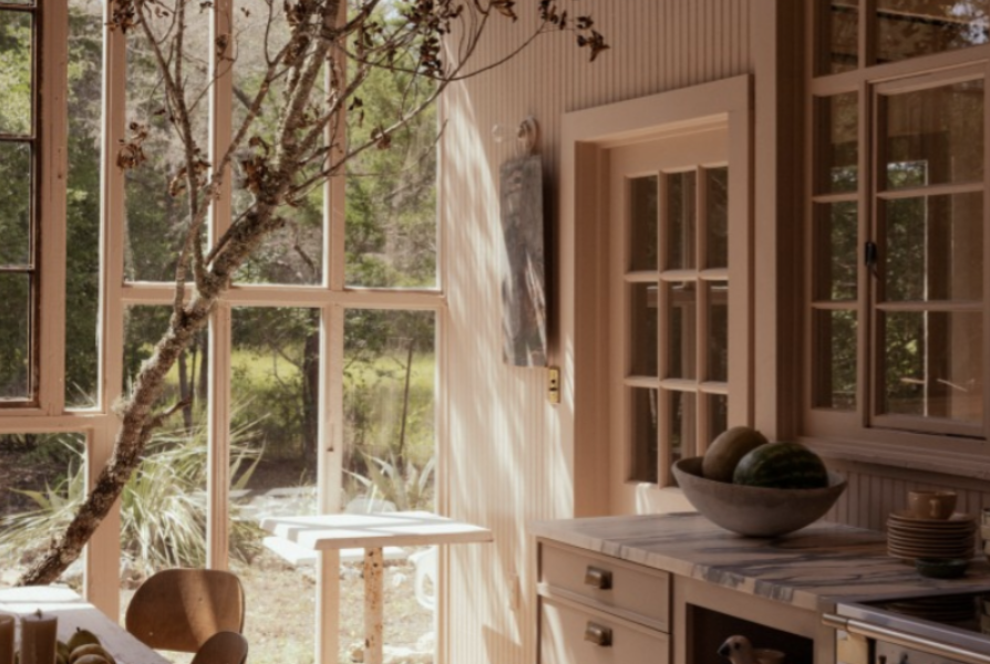 Interior of a sunlit room with large windows, wooden furniture, and a bowl of melons on the counter.