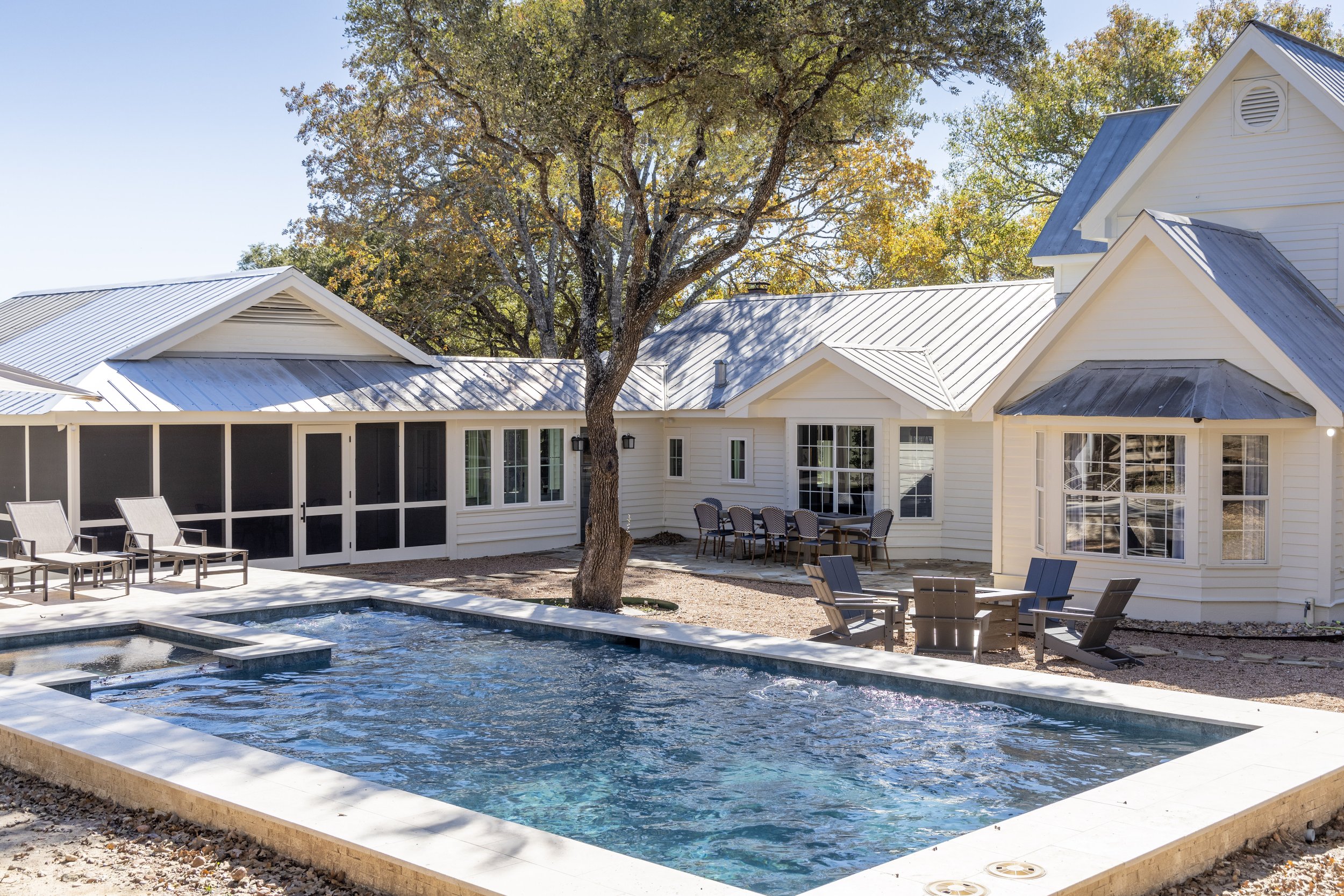 Backyard scene with a swimming pool, outdoor chairs, a table with chairs, a large tree, and a white house with a metal roof.