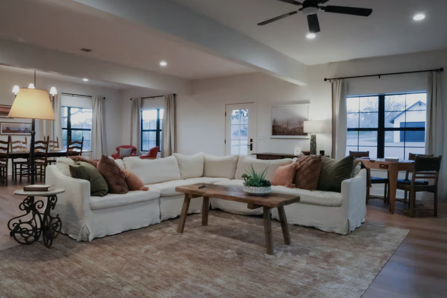A spacious living room with a large white sectional sofa decorated with assorted pillows and a wooden coffee table with a plant. The room features hardwood floors, large windows with white curtains, and is illuminated by ceiling lights, a floor lamp, and a table lamp. Additional furniture includes a dining table with chairs and a small desk or side table.