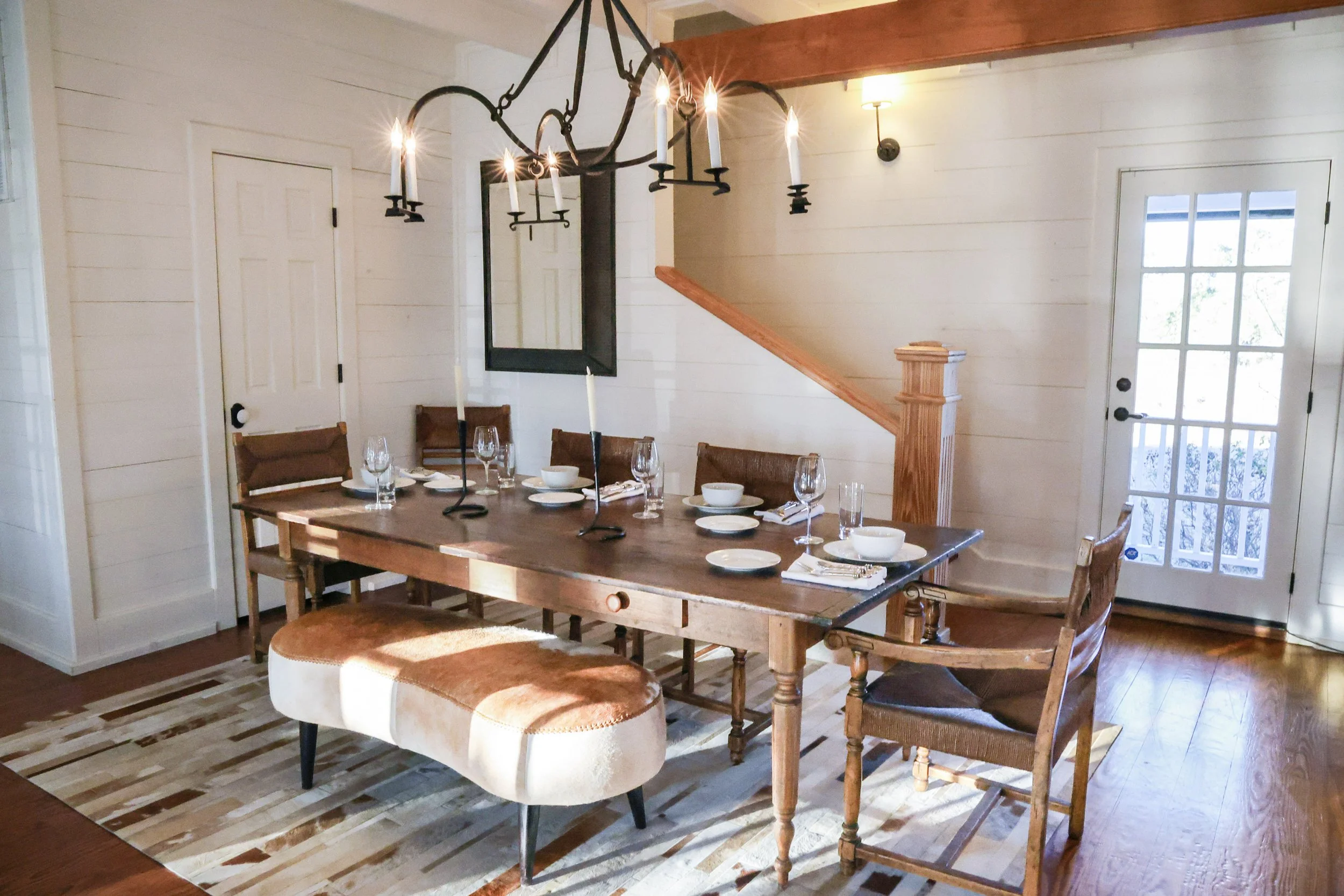 A dining room with a wooden table set with plates, glasses, and bowls, surrounded by wooden chairs and an upholstered bench. A chandelier hangs above the table, and a mirror and wall lamps are on the cream-colored walls. There's a door with glass panels and a staircase in the background.