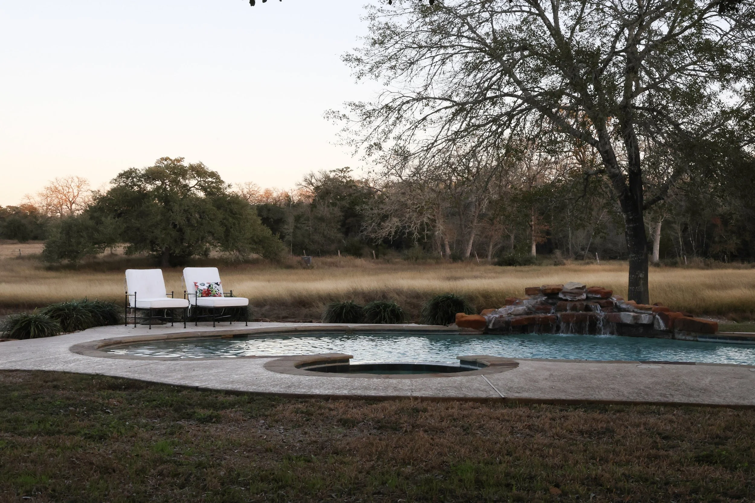 A backyard with a swimming pool, surrounded by grass and trees, two white chairs with cushions, and a large tree to the right, located in a rural area with open fields and a sunset sky.