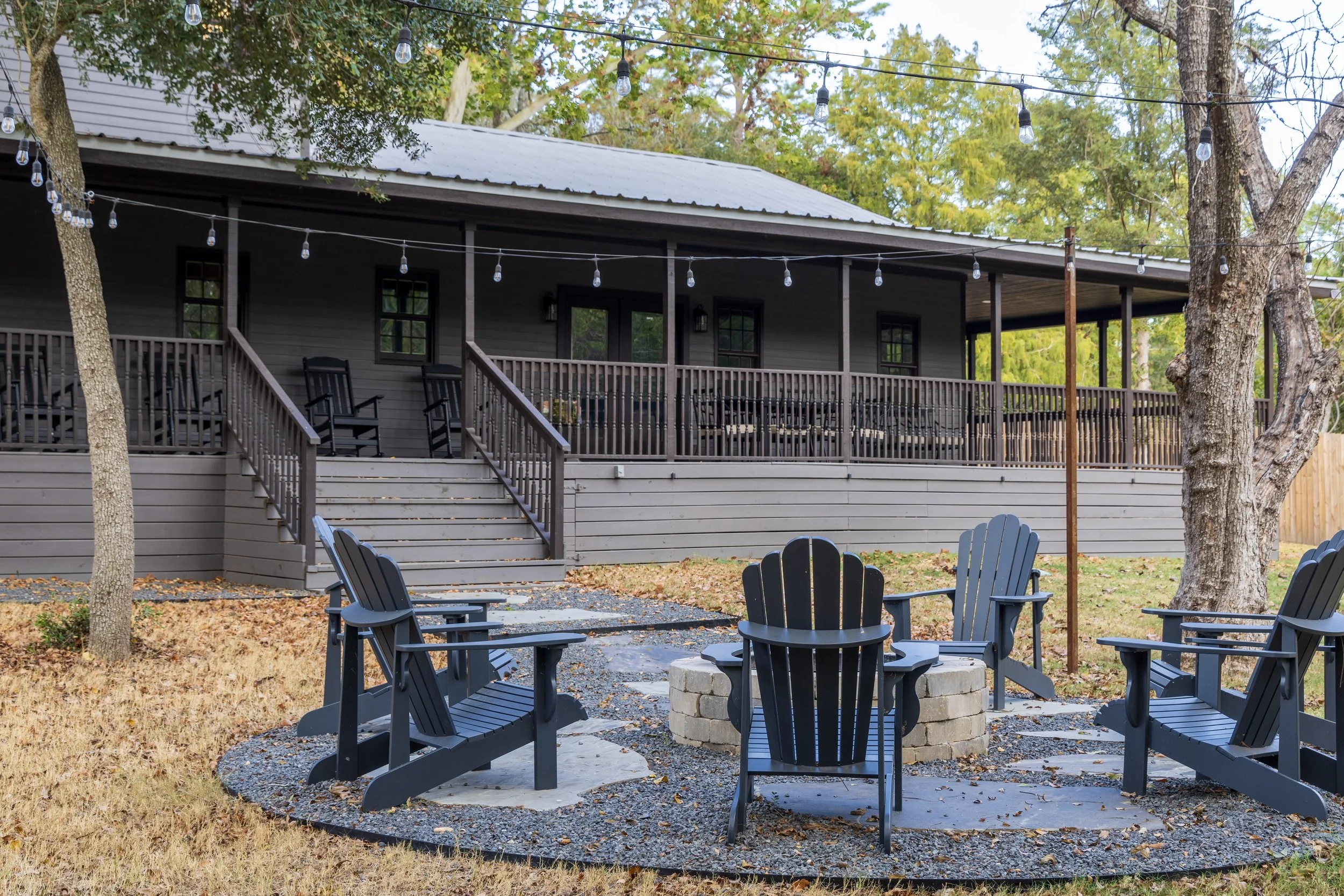 A backyard patio with a fire pit surrounded by black Adirondack chairs, trees, and an elevated porch with string lights