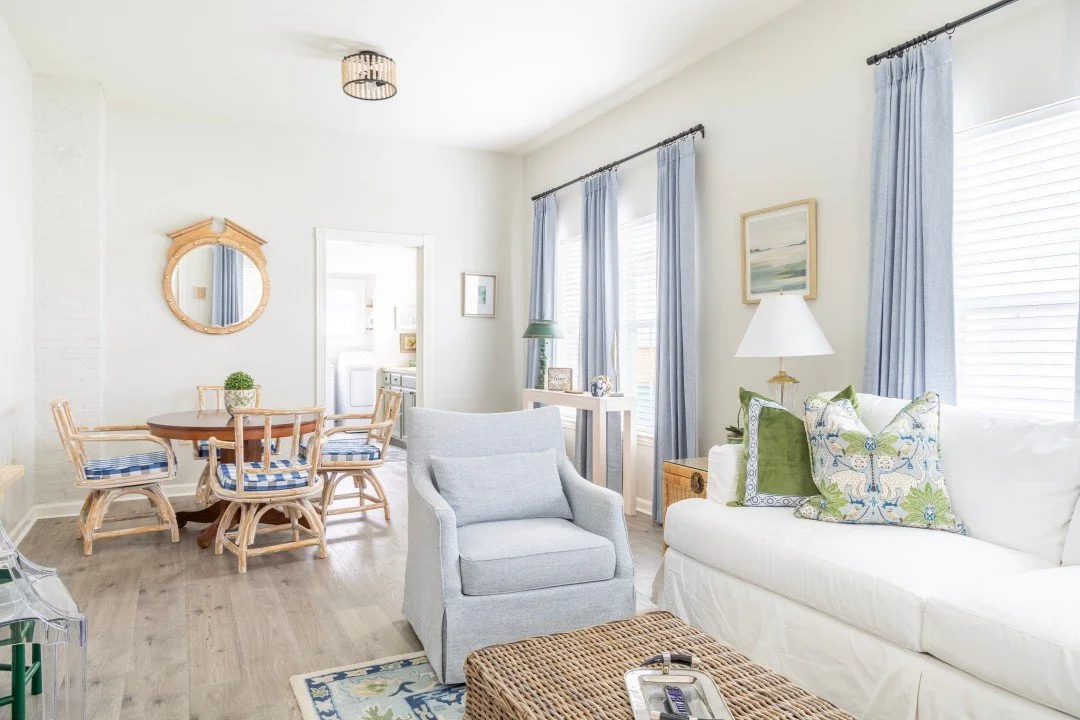 Bright living room with white walls, light wood flooring, a white sofa with patterned pillows, a light gray armchair, and a wicker coffee table. There are three large windows with blue curtains, a framed picture beside the windows, and a table lamp. The dining area features a round wooden table with six matching chairs with blue and white striped cushions. A mirror hangs on the wall above the dining table.