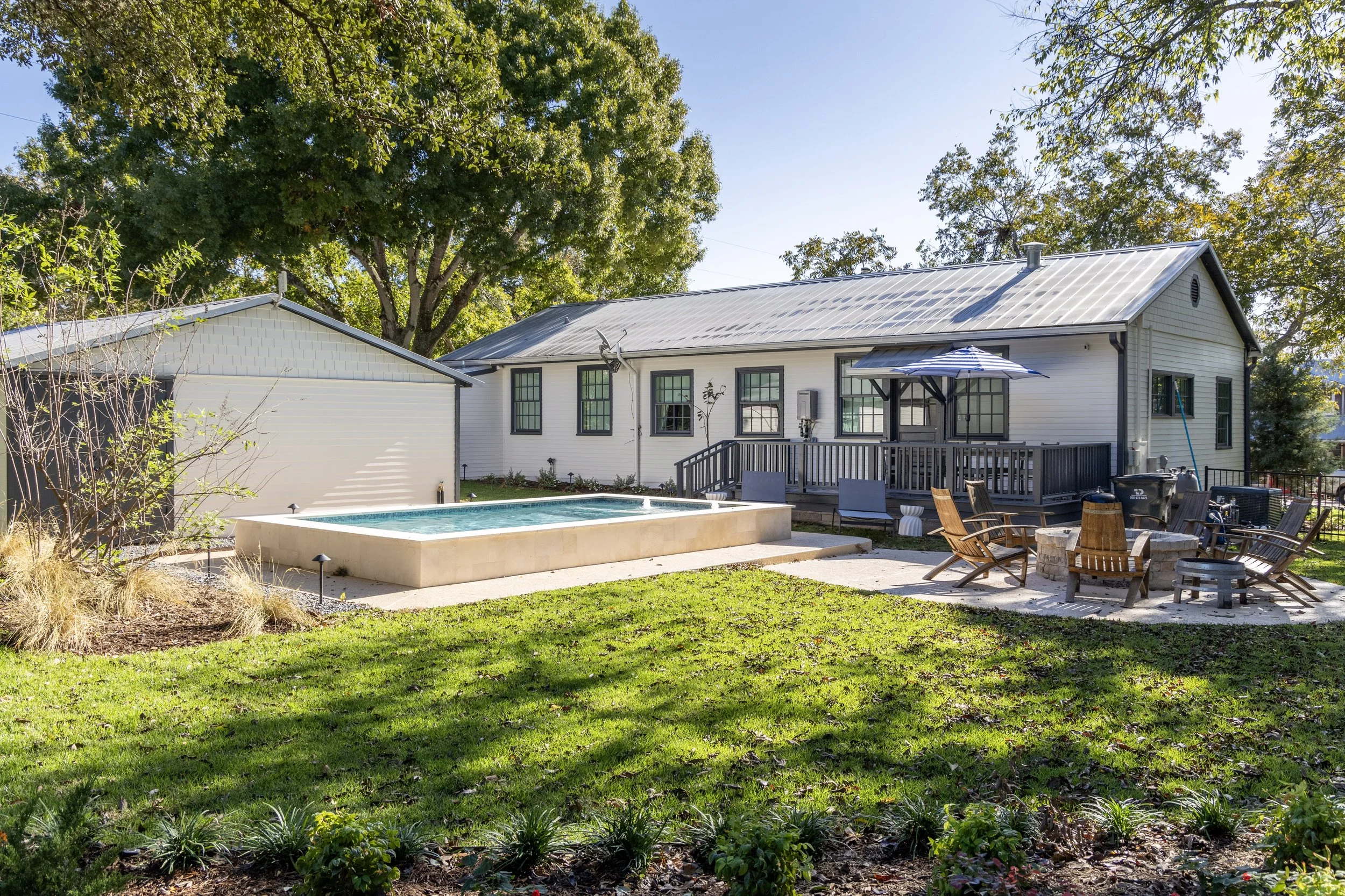 Backyard with a pool, outdoor chairs, and a patio, enclosed by trees and a house with white siding and multiple windows.