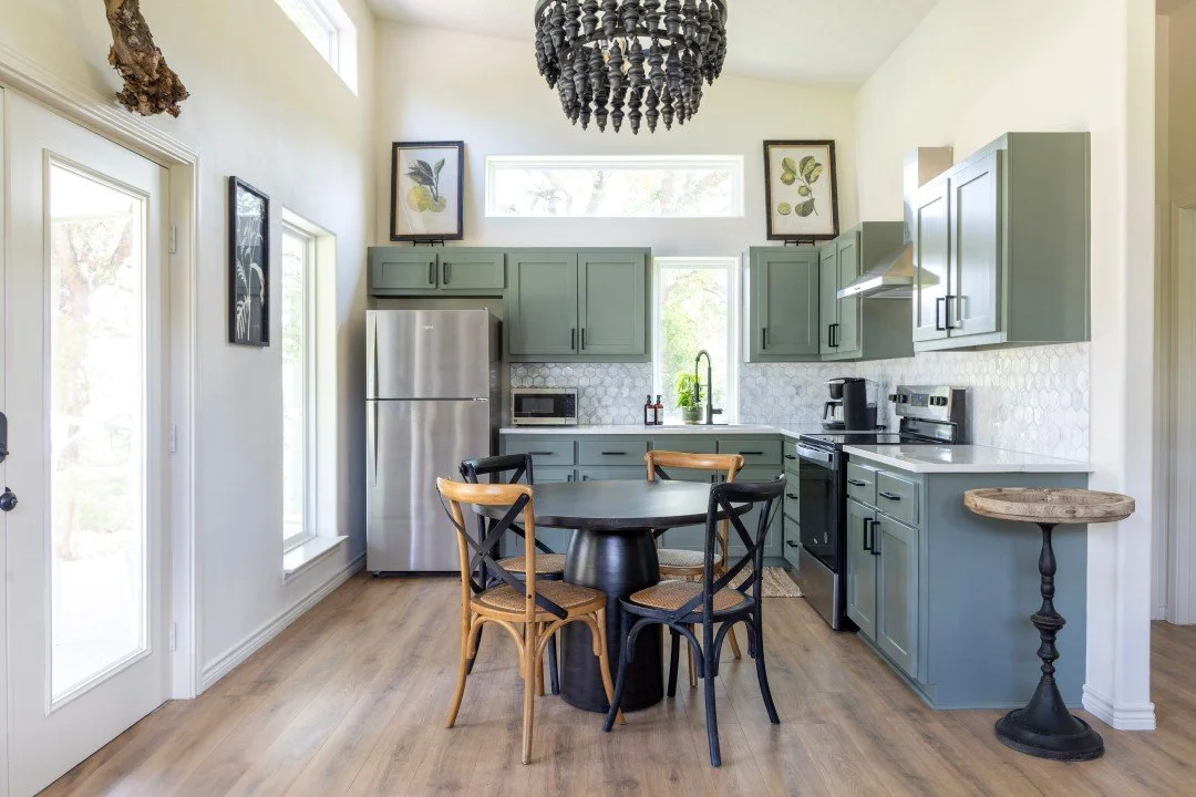 Modern kitchen with sage green cabinets, stainless steel refrigerator, black stove, and small round dining table with mixed black and natural wood chairs. Decorative artwork and framed botanical prints on walls, chandelier hanging from ceiling, and wooden floor.