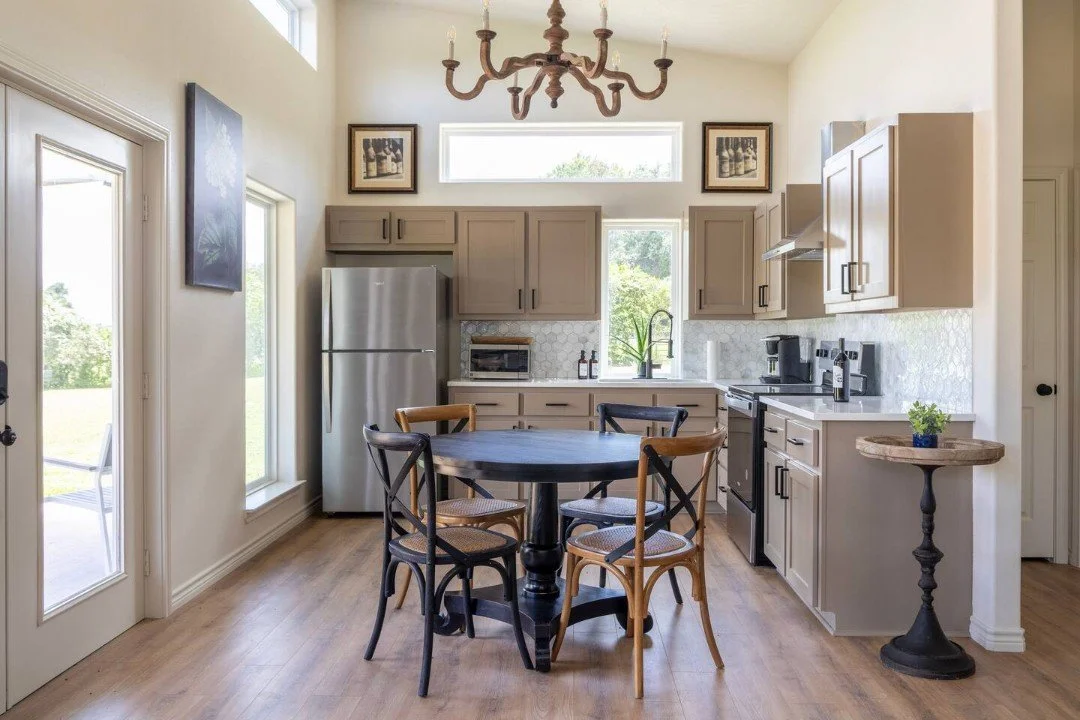 Modern kitchen with beige cabinets, stainless steel refrigerator, black stove, small round dining table with four chairs, wooden flooring, chandelier, and natural light from windows.