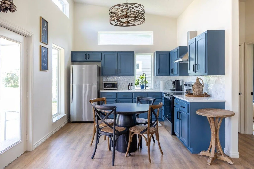 A modern kitchen with blue cabinets, a round dining table with four chairs, stainless steel refrigerator, black stove, and light wood flooring.