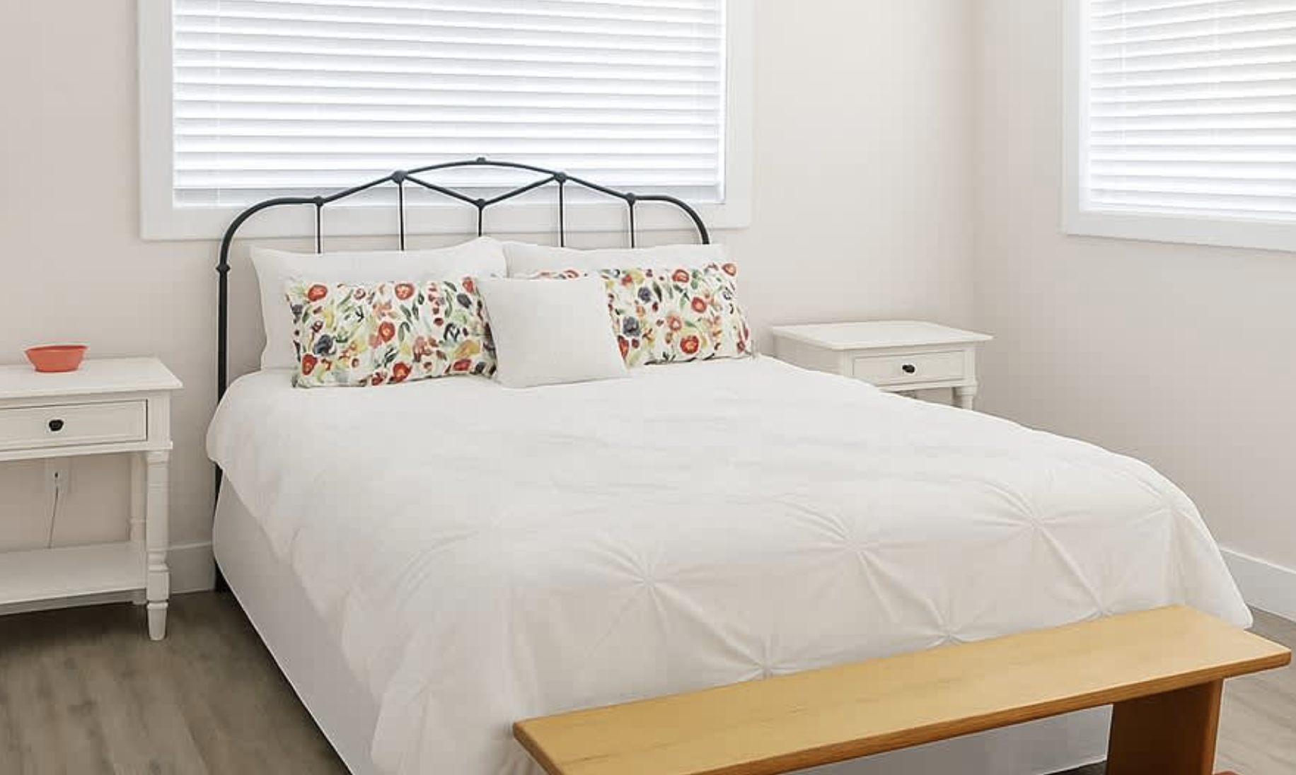 A neatly made bed with a white duvet, floral pillows, and a white pillow, flanked by white nightstands with small drawers, in a bright room with white walls and blinds.