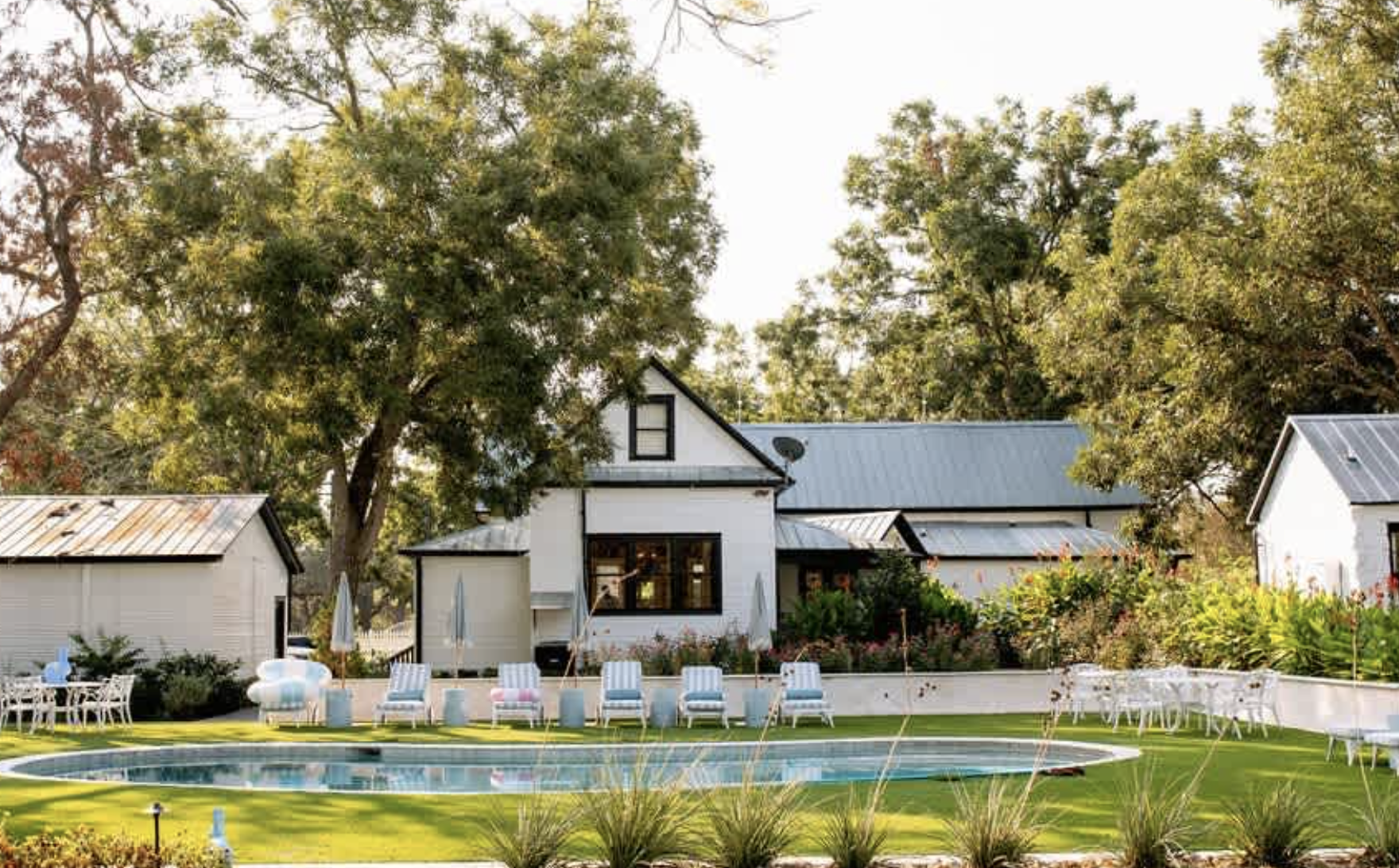 A backyard with a swimming pool, white loungers, tables, and chairs, surrounded by green grass, bushes, and tall trees near a white house with a metal roof.