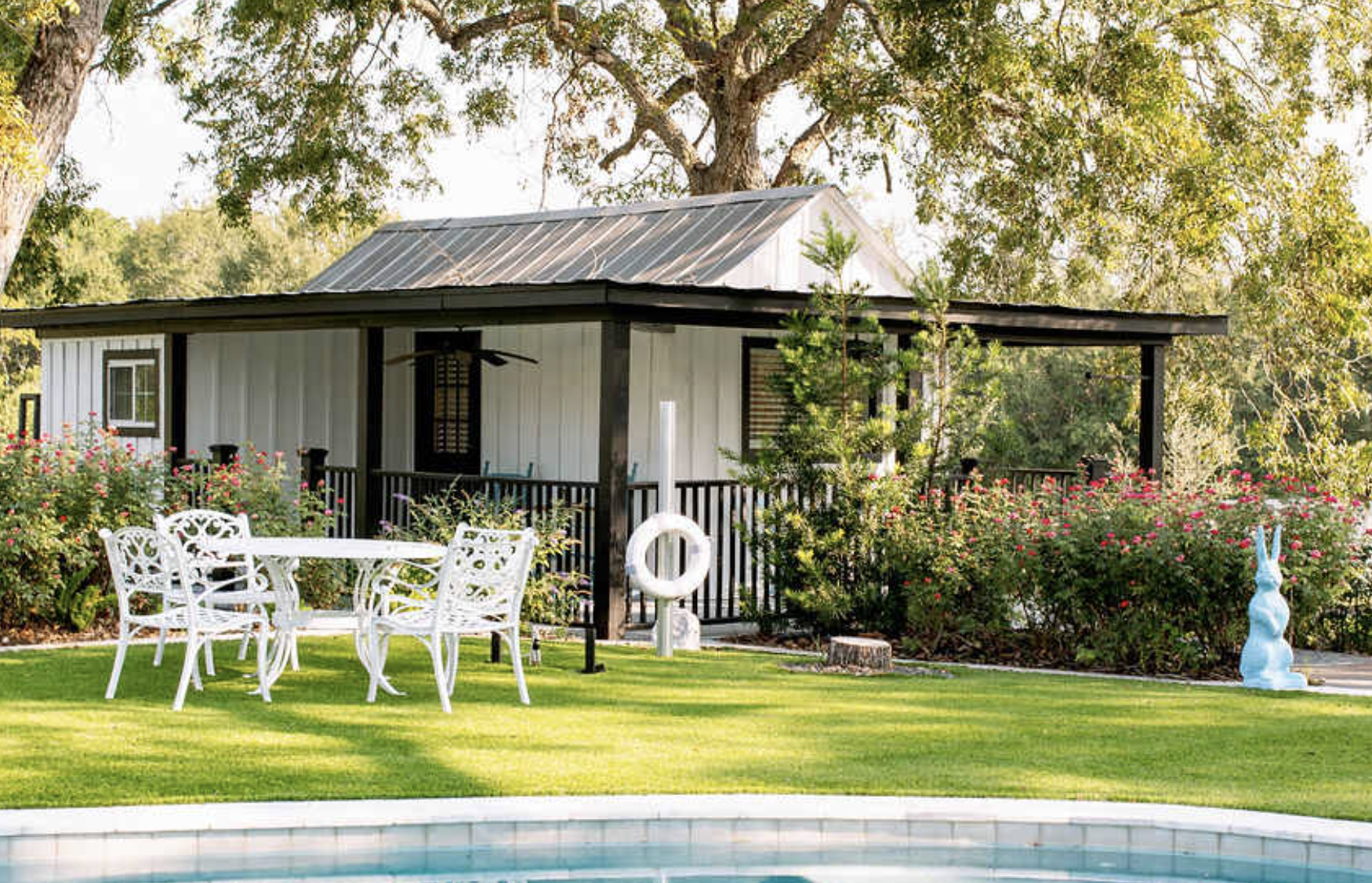 A backyard with a swimming pool, white patio table with chairs, lush green grass, blooming bushes, and a small white house with a porch and black railing, surrounded by tall trees.