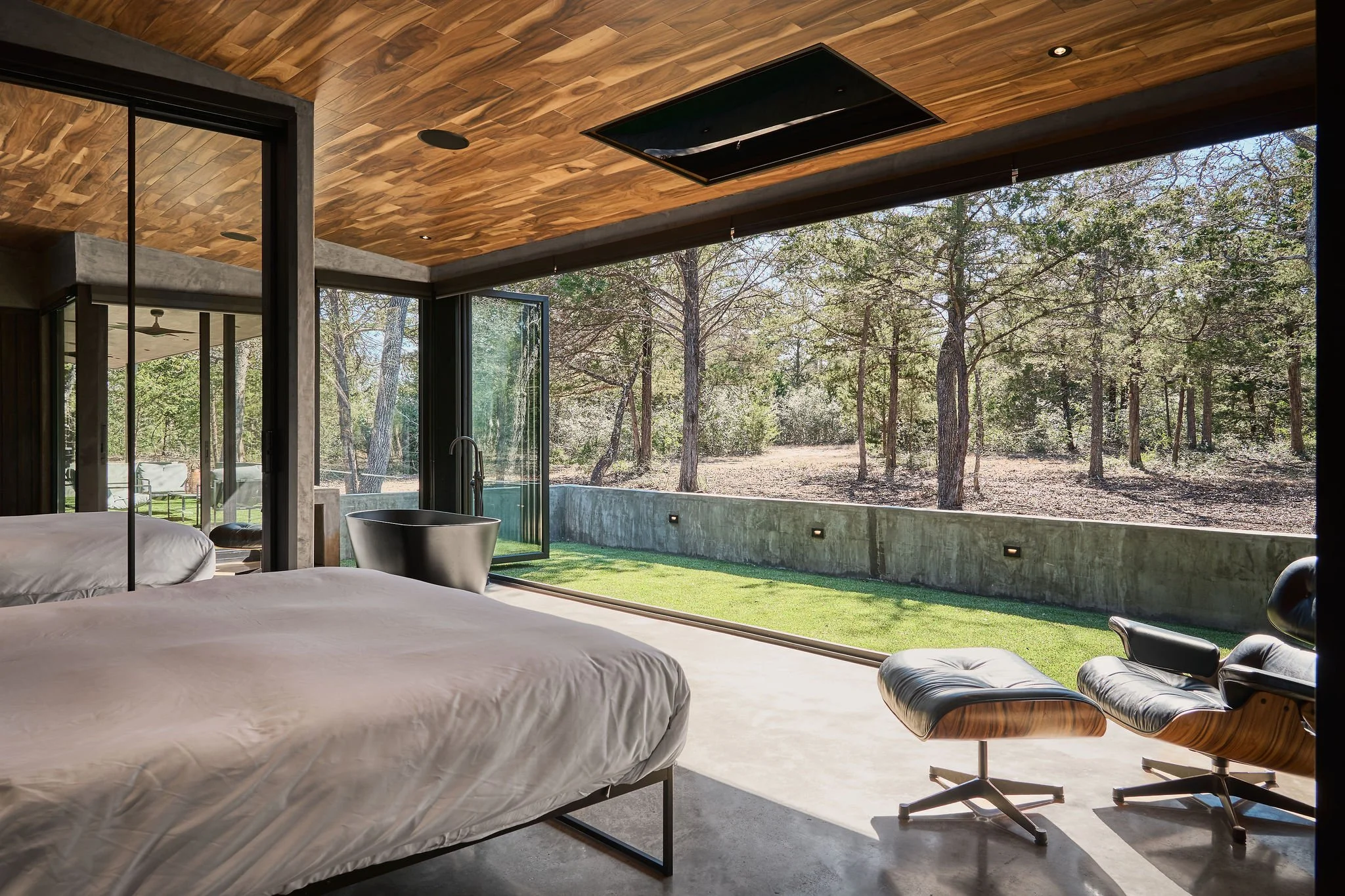 Modern bedroom with large glass sliding doors opening to a wooded outdoor area, featuring two beds, a black bathtub, a lounge chair, and a wooden ceiling.