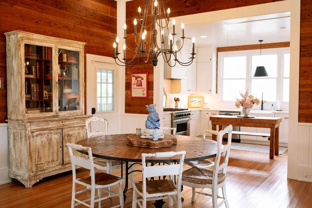 A cozy dining area with a round wooden table surrounded by white chairs, a large rustic chandelier hanging overhead, and a wooden china cabinet on a wood-paneled wall. In the background, a bright kitchen with white cabinets, a window with flowers, and a wooden kitchen island are visible.