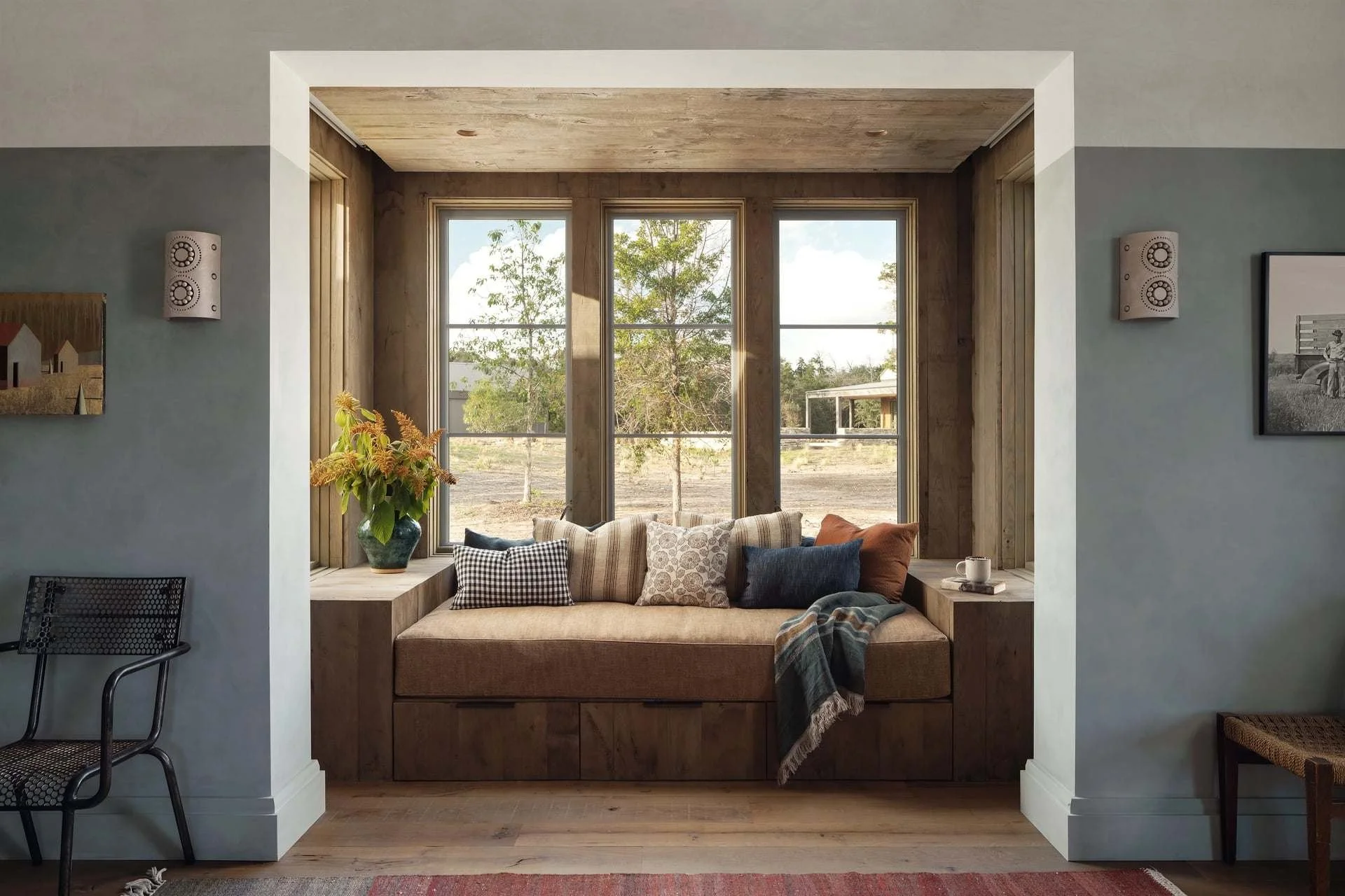 A cozy window nook with a built-in wooden bench, cushions, and a blanket, overlooking a rural landscape with trees and a modern house in the distance.