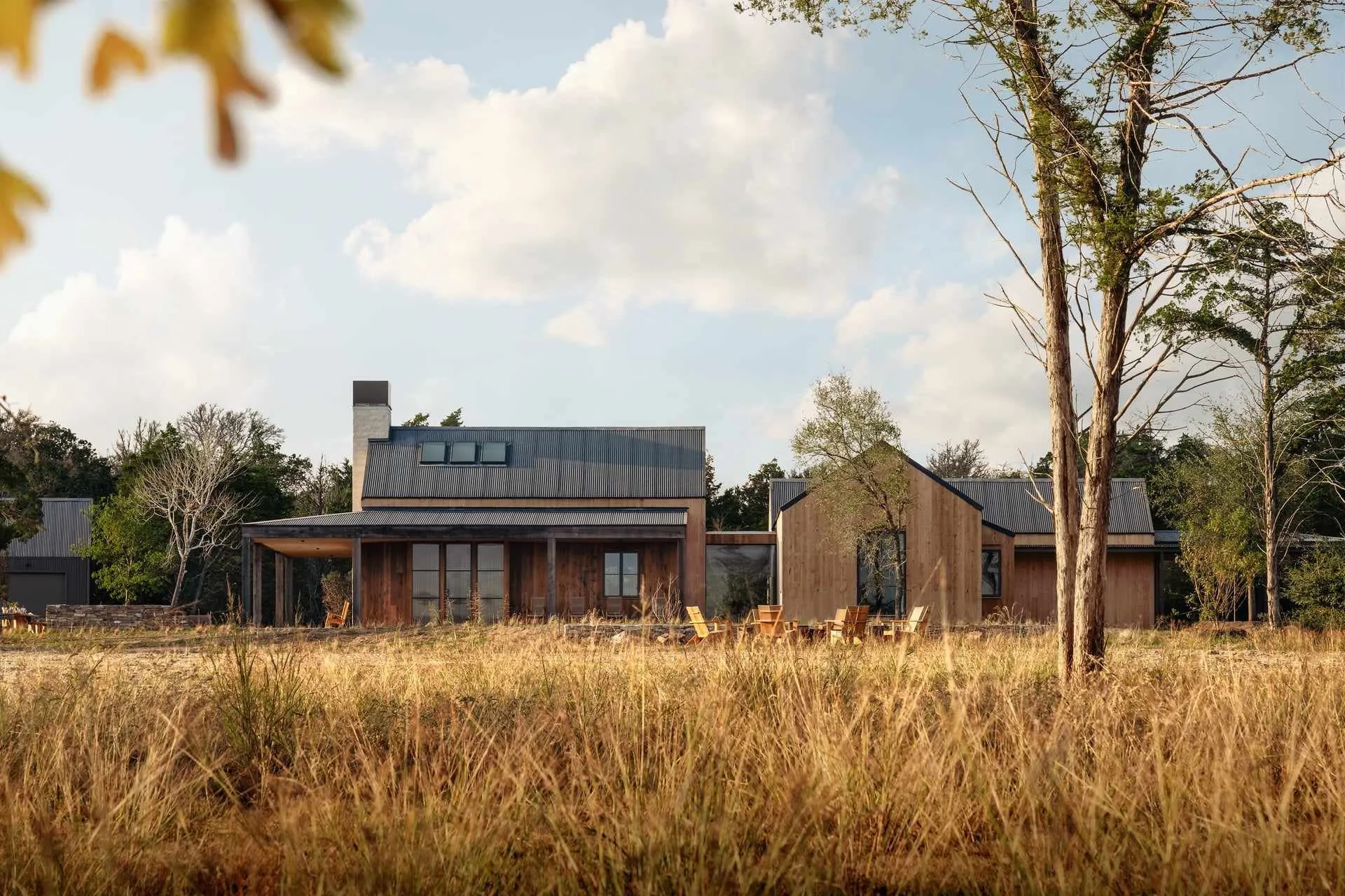 Modern house with wooden exterior and dark metal roof, surrounded by trees and tall grass, with outdoor seating on patio, under a partly cloudy sky.