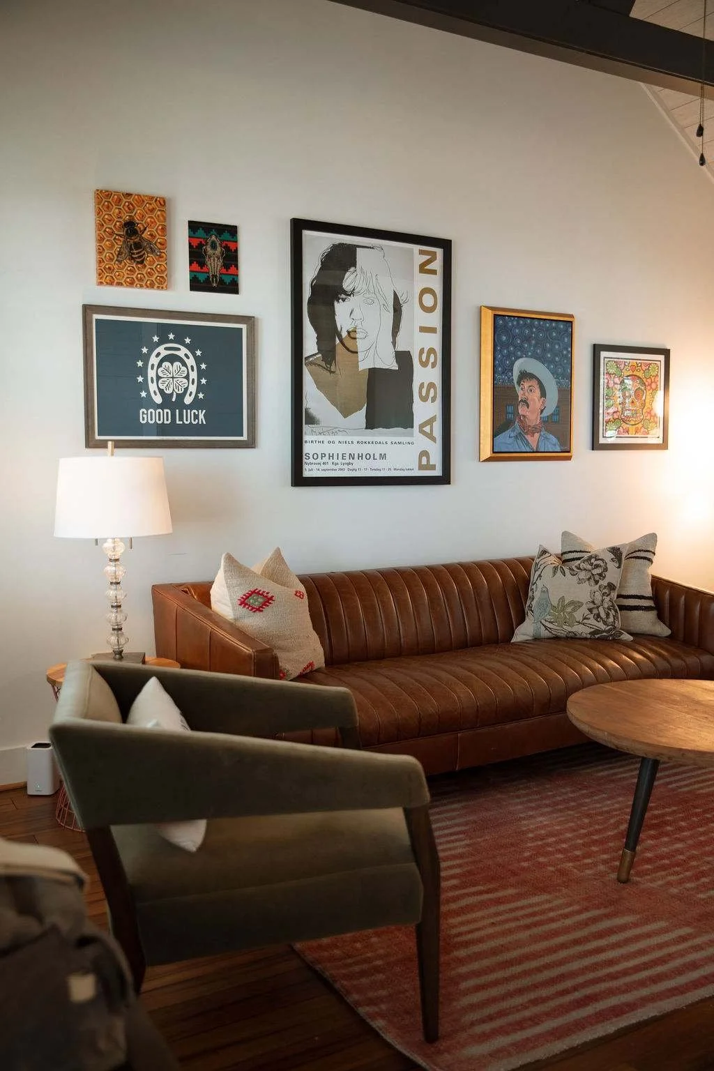 Living room with a brown leather sofa, throw pillows, a wooden coffee table, a gray armchair with a pillow, and wall art including a poster, framed artwork, and a photo, with a white wall and lamps.