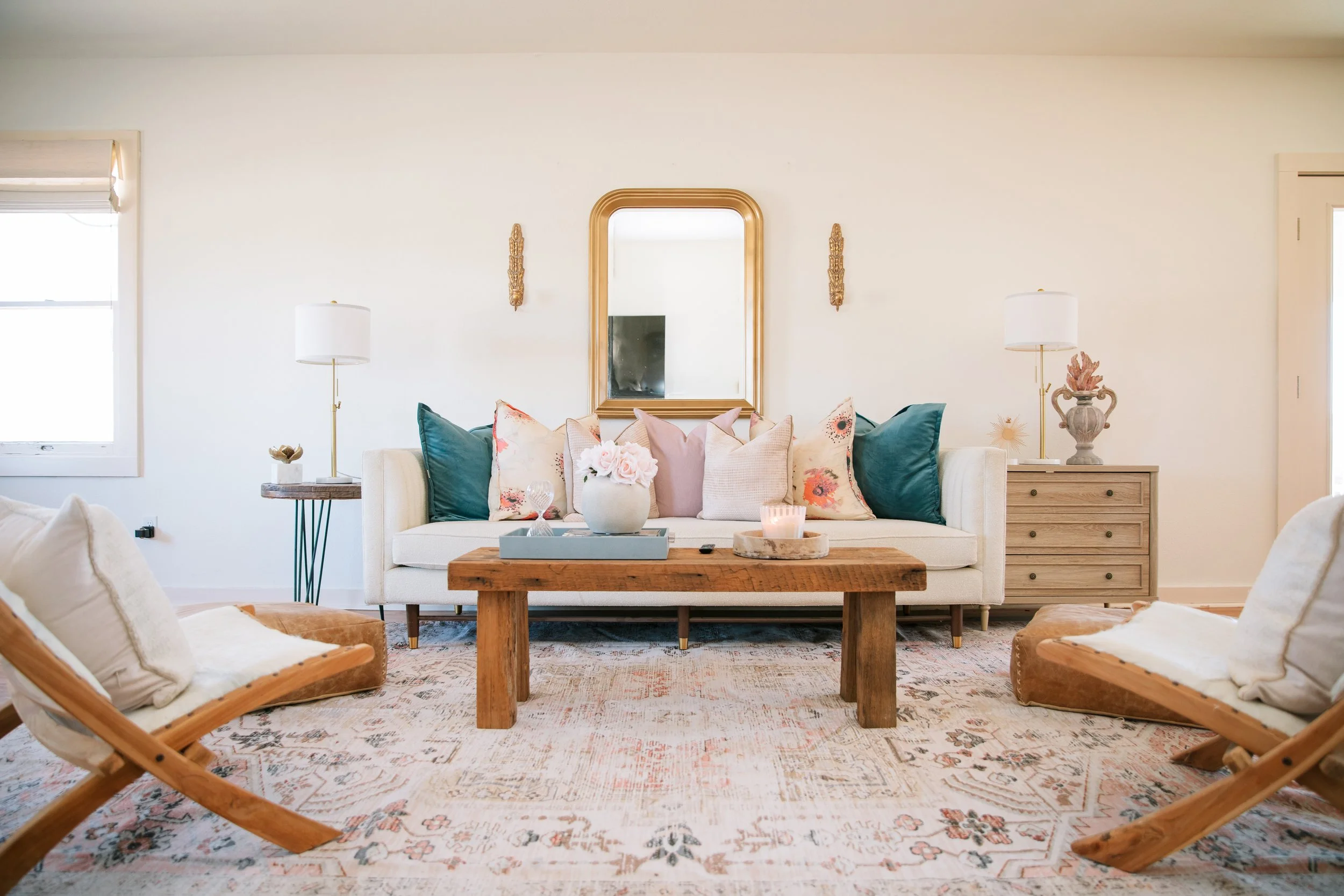 Bright living room with a white sofa adorned with colorful throw pillows, a wooden coffee table, and two wooden chairs with cushions. A large mirror hangs above the sofa, flanked by two wall sconces. There are two side tables with lamps and decor, and a window to the left allowing natural light. The room has a neutral wall color and an oriental-style area rug.