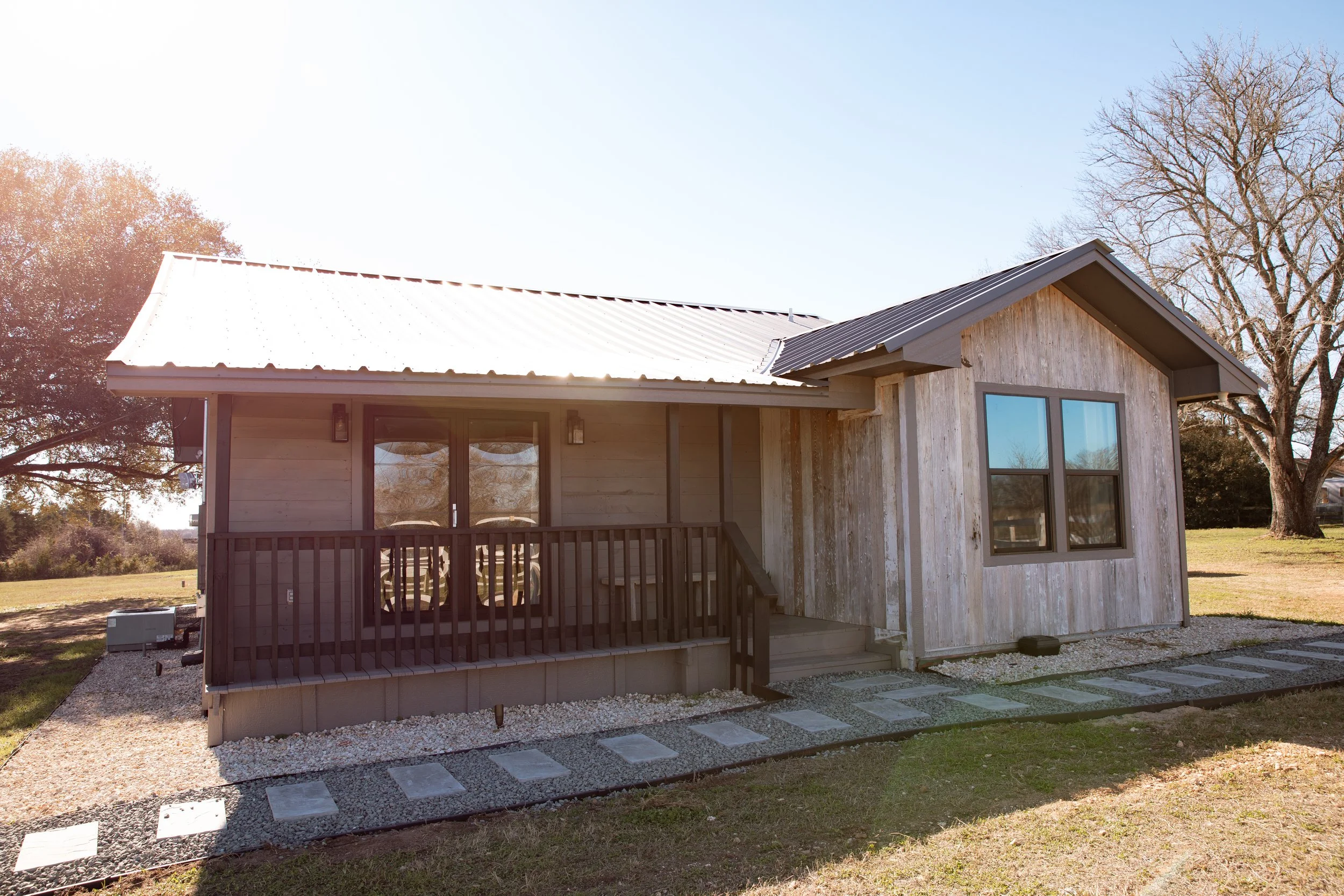 Front view of a small wooden house with a porch and gray window frames, surrounded by a lawn and trees.