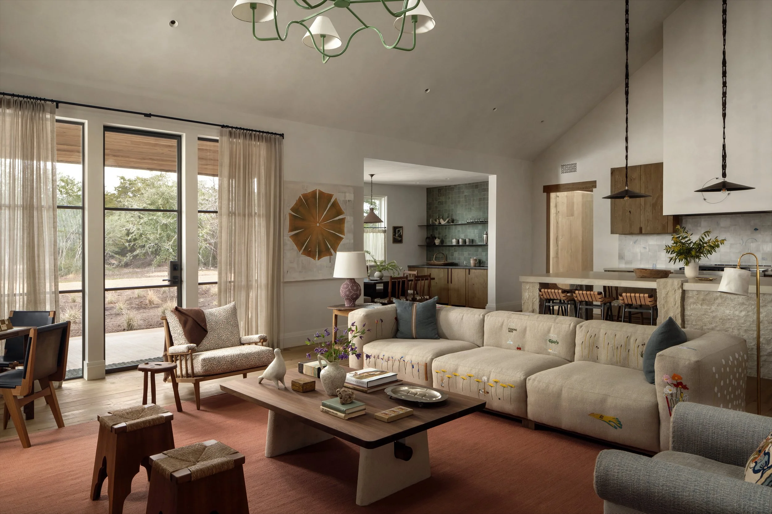 Living room with cream sectional sofa, wooden coffee table, armchair, and pink rug. Large windows with sheer curtains showing outdoor greenery. Open kitchen with bar stools, shelves, and wood accents.