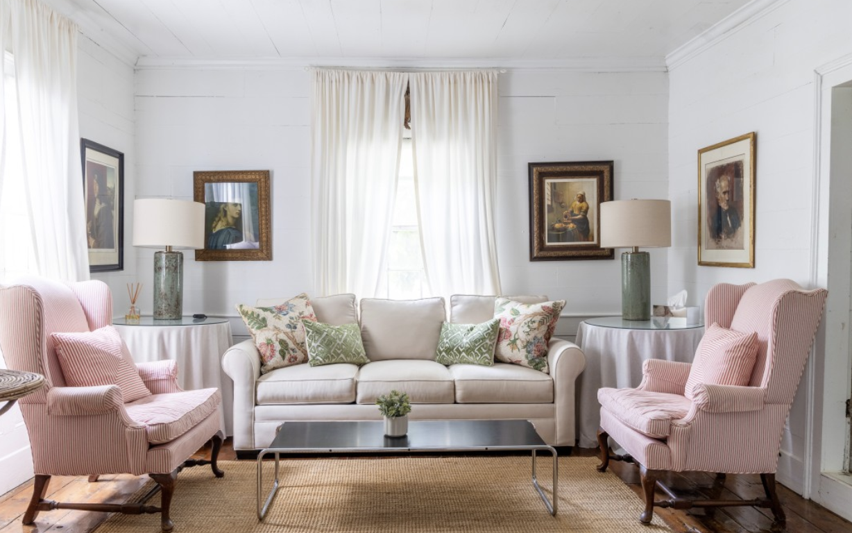 Living room with white walls, three framed paintings, white curtains, a white sofa with throw pillows, two pink armchairs, and side tables with lamps.
